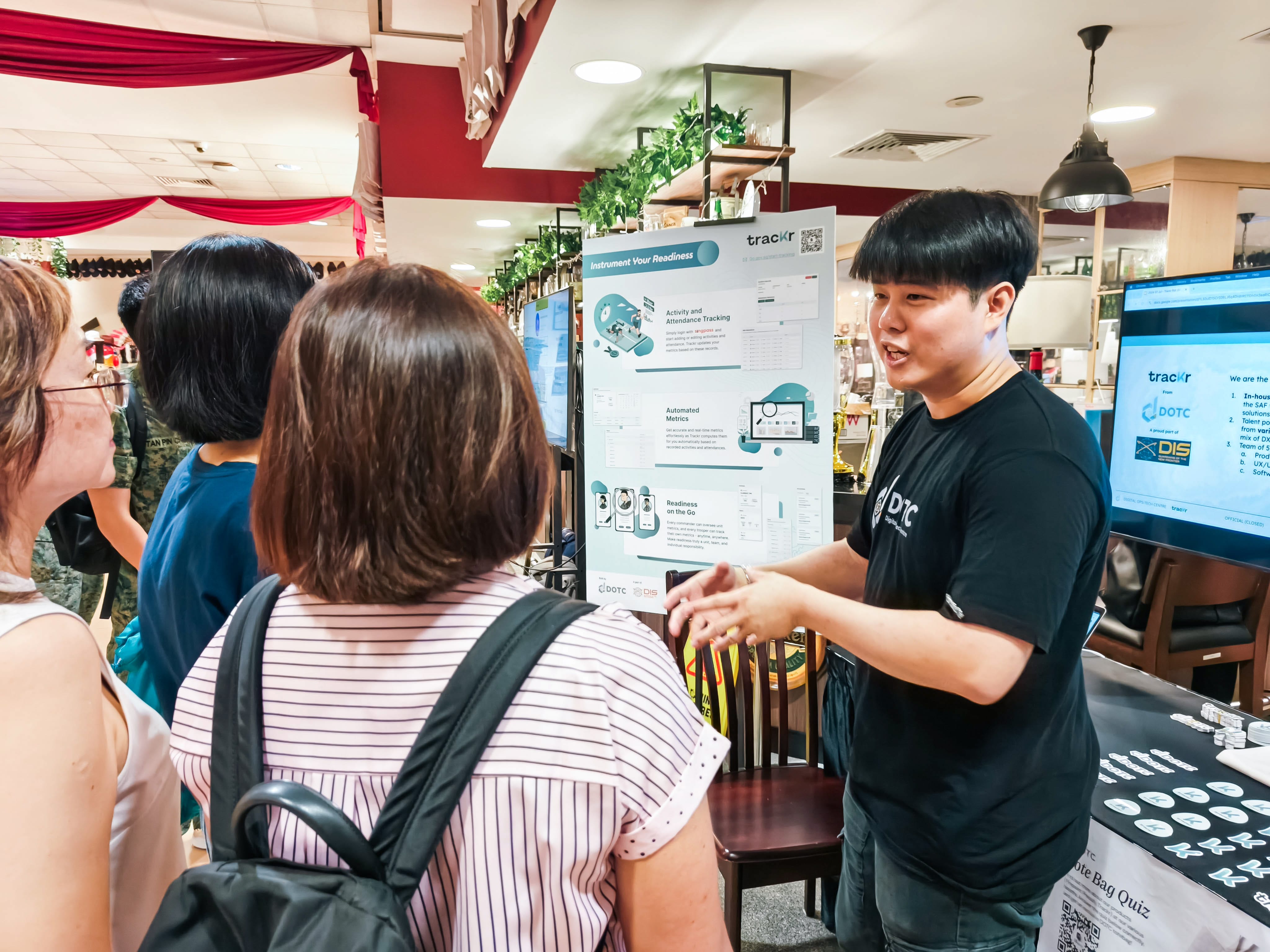 A young man in a black DTC shirt presents a Trackr poster to a small group of attendees at an indoor event.