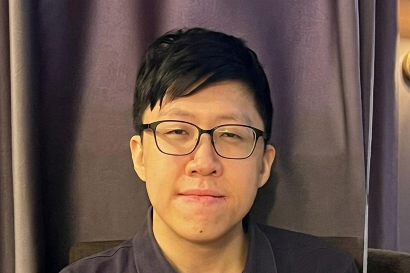 Young man with black rectangular glasses and short dark hair wearing a dark polo shirt, seated near a purple curtain.
