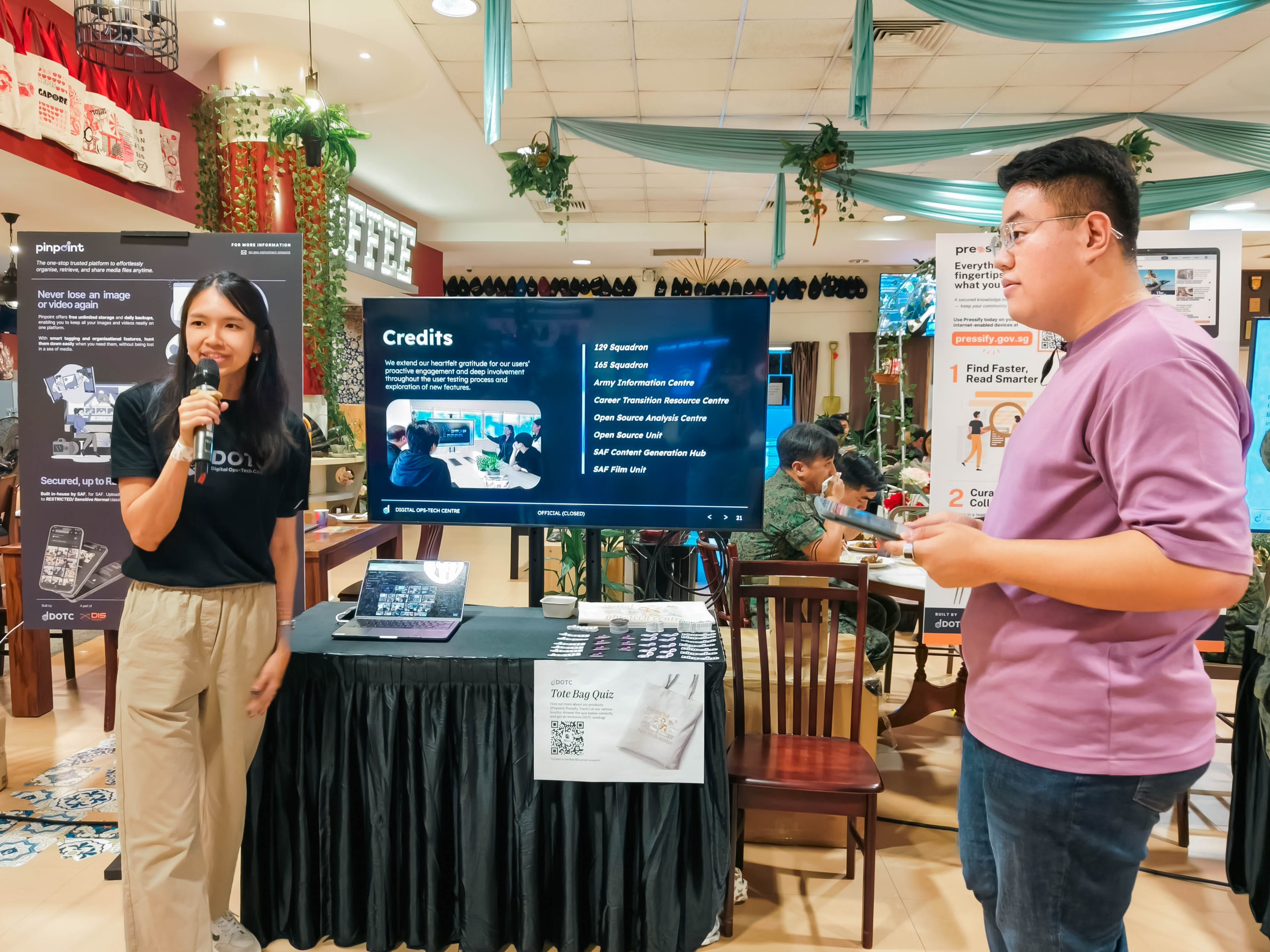 Two presenters stand beside a screen showing a Credits slide at an indoor event with display banners nearby.