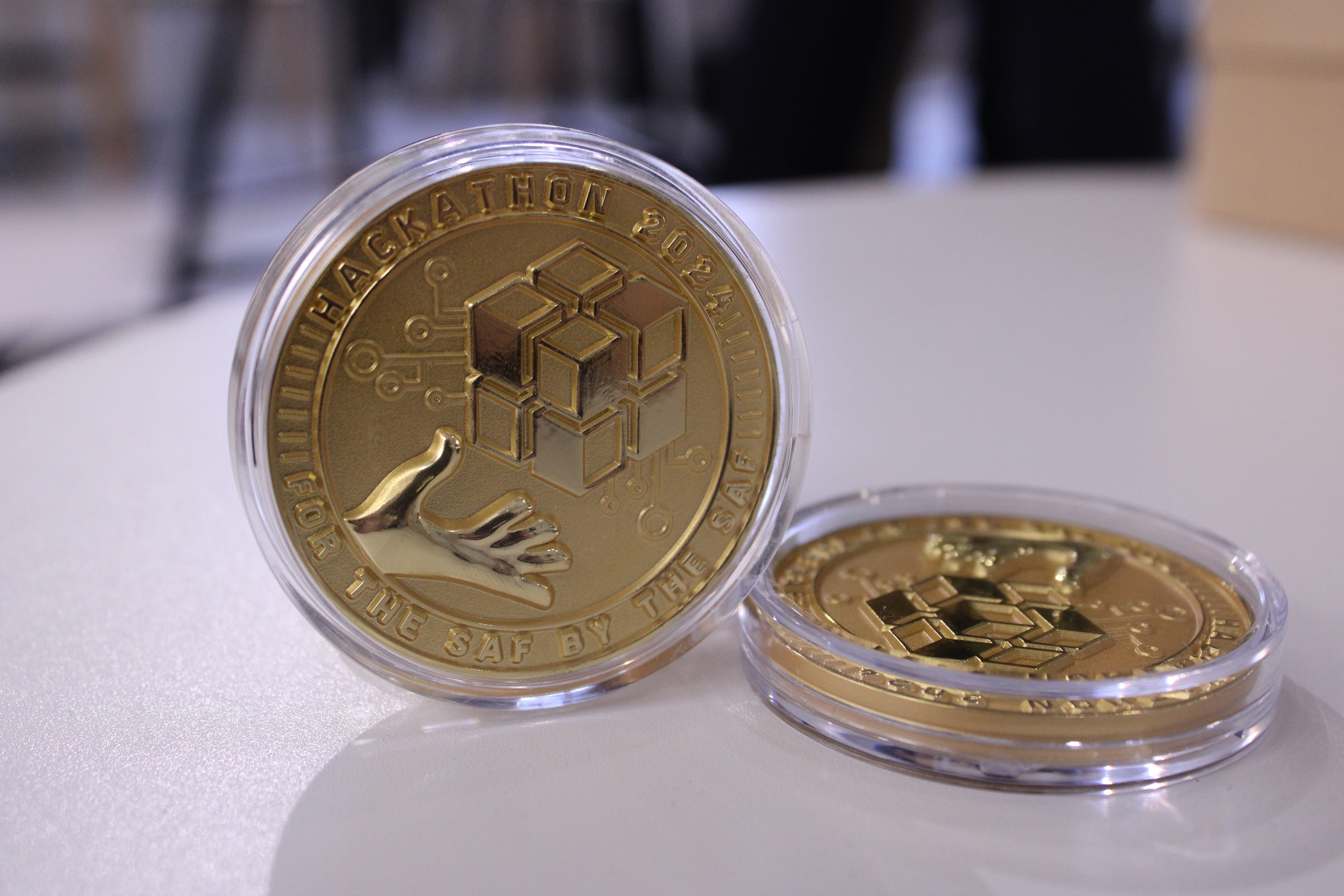 Two gold coins in clear cases; one reads Hackathon 2024 and shows cubes and hands on a table.