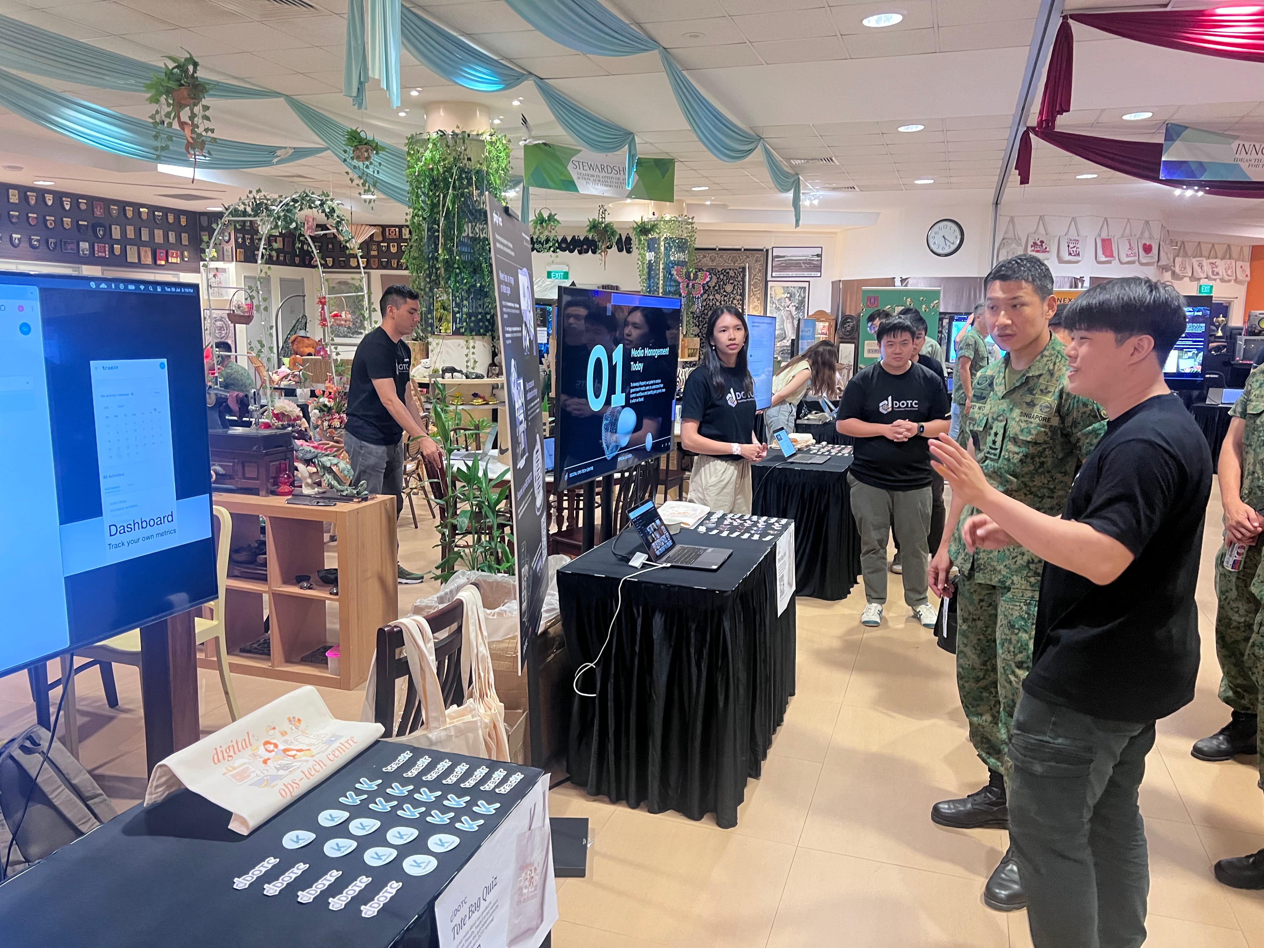 People at an indoor tech exhibition with display screens, booths, and a Singapore military officer speaking with attendees.