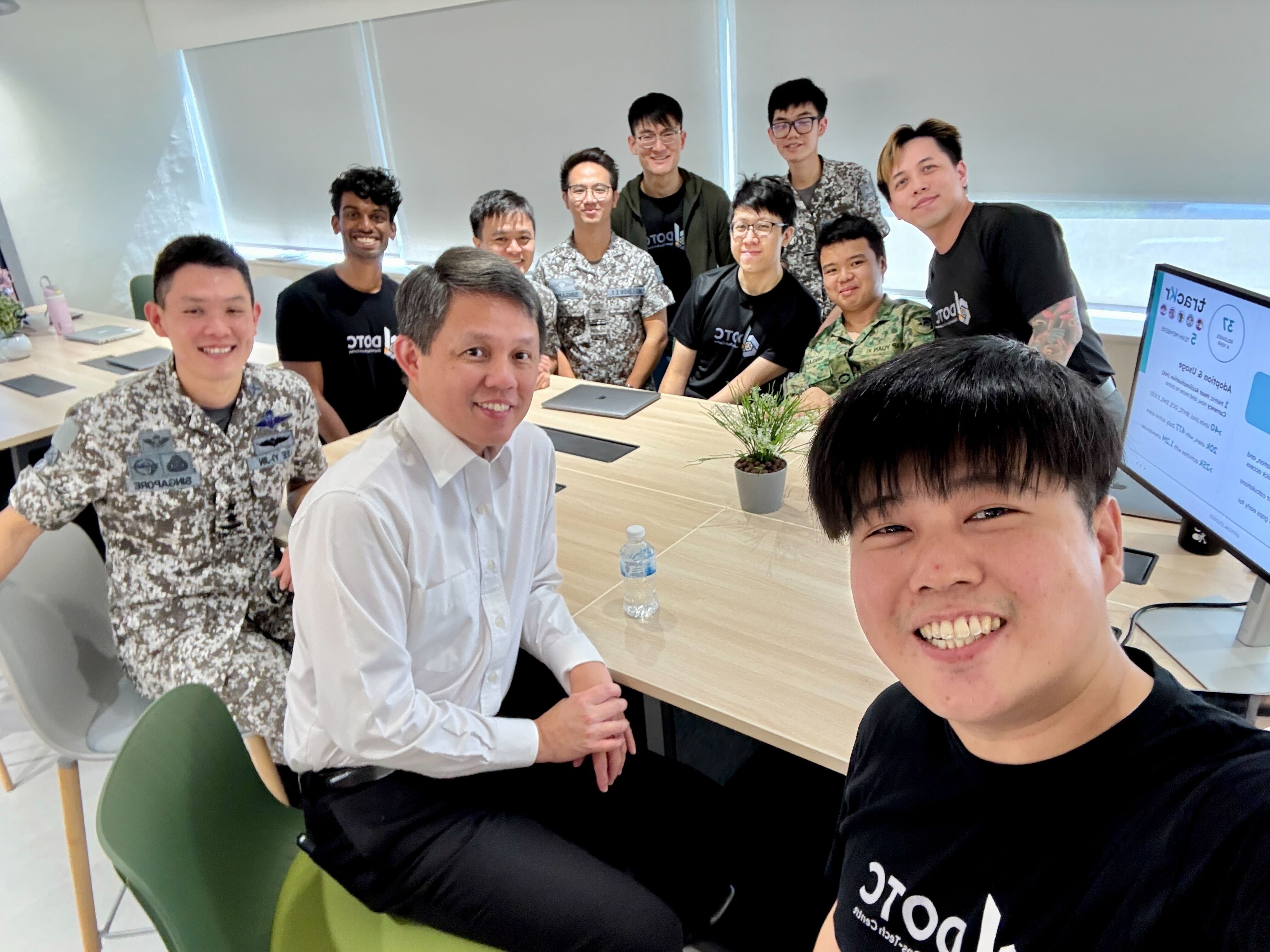 A group of about eleven people smile for a selfie around a wooden table in a bright modern office.