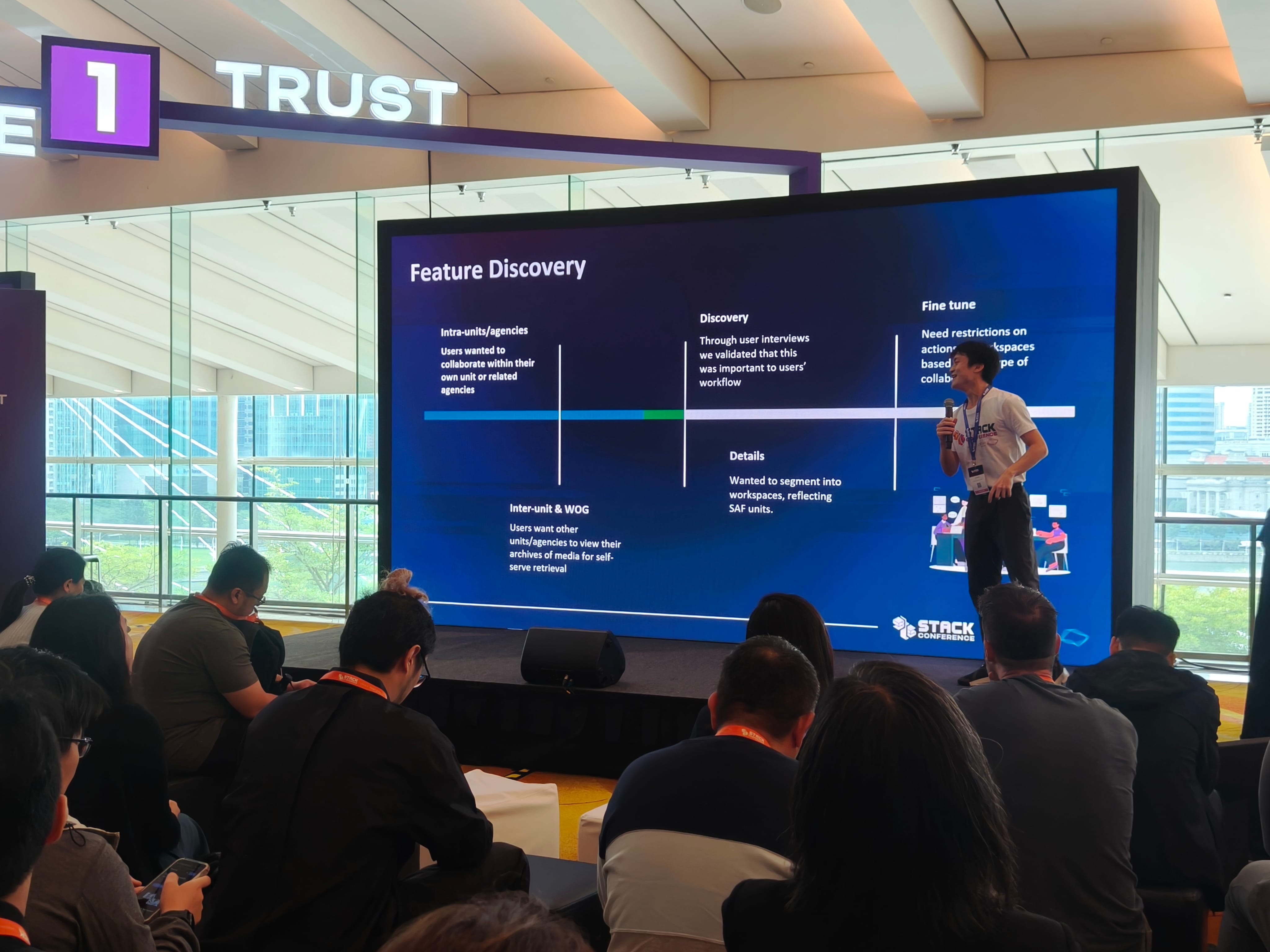 A speaker holds a mic on stage at Stack Conference presenting a Feature Discovery slide to a seated audience.