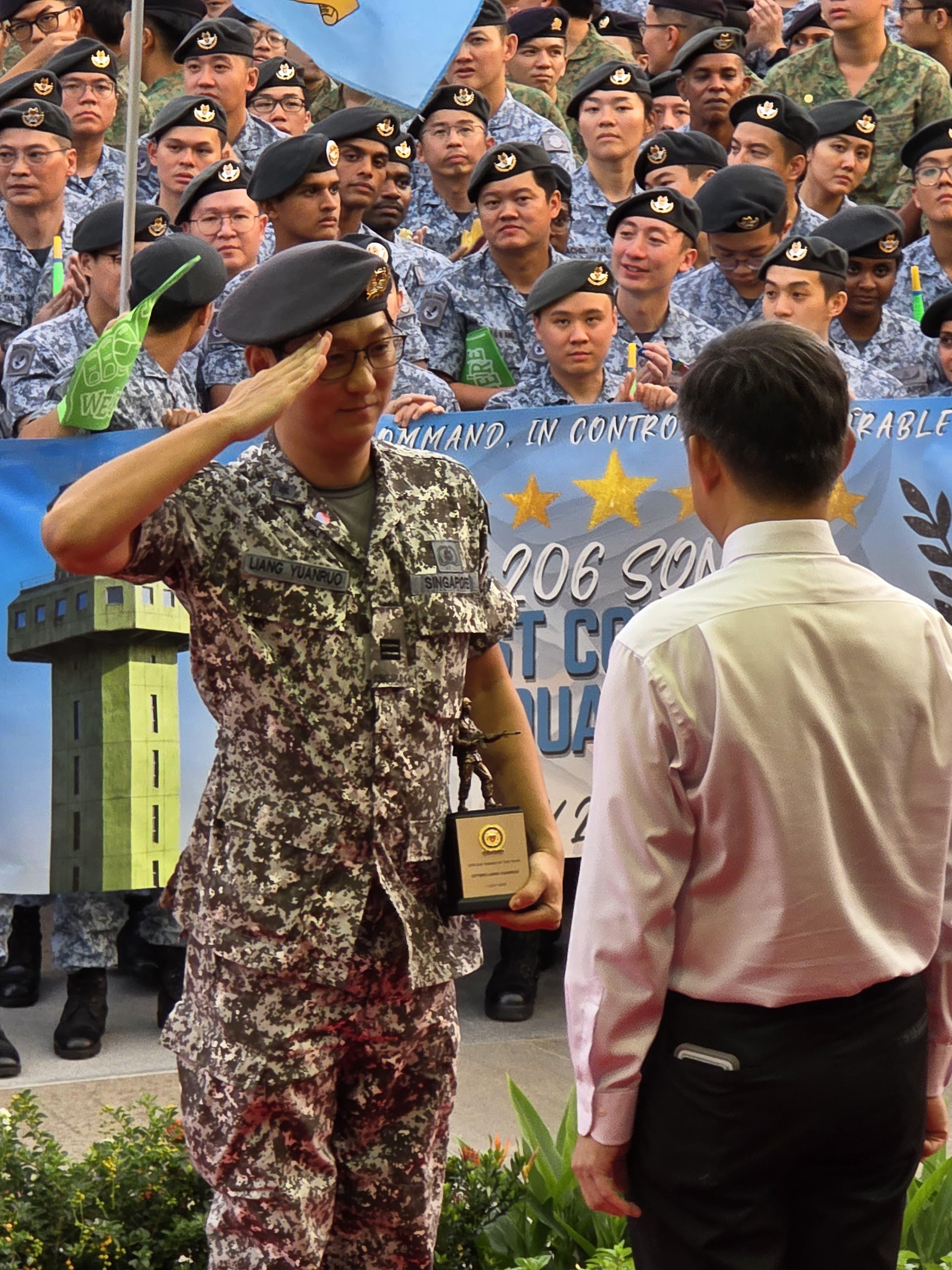 A medium shot of an East Asian man in a pixelated Singapore military camouflage uniform and black beret, holding a small trophy while rendering a formal salute to an official in a white shirt, with a crowd of soldiers in uniform standing in the background.