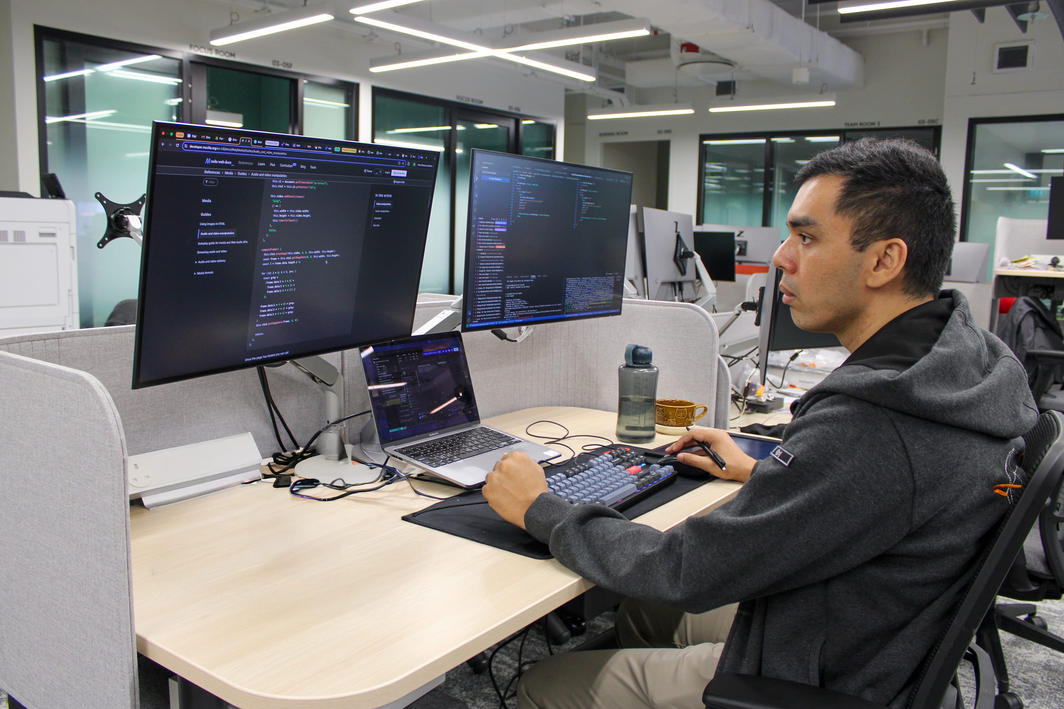 Person at desk in open office using two large monitors and a laptop showing code, hands on keyboard and mouse