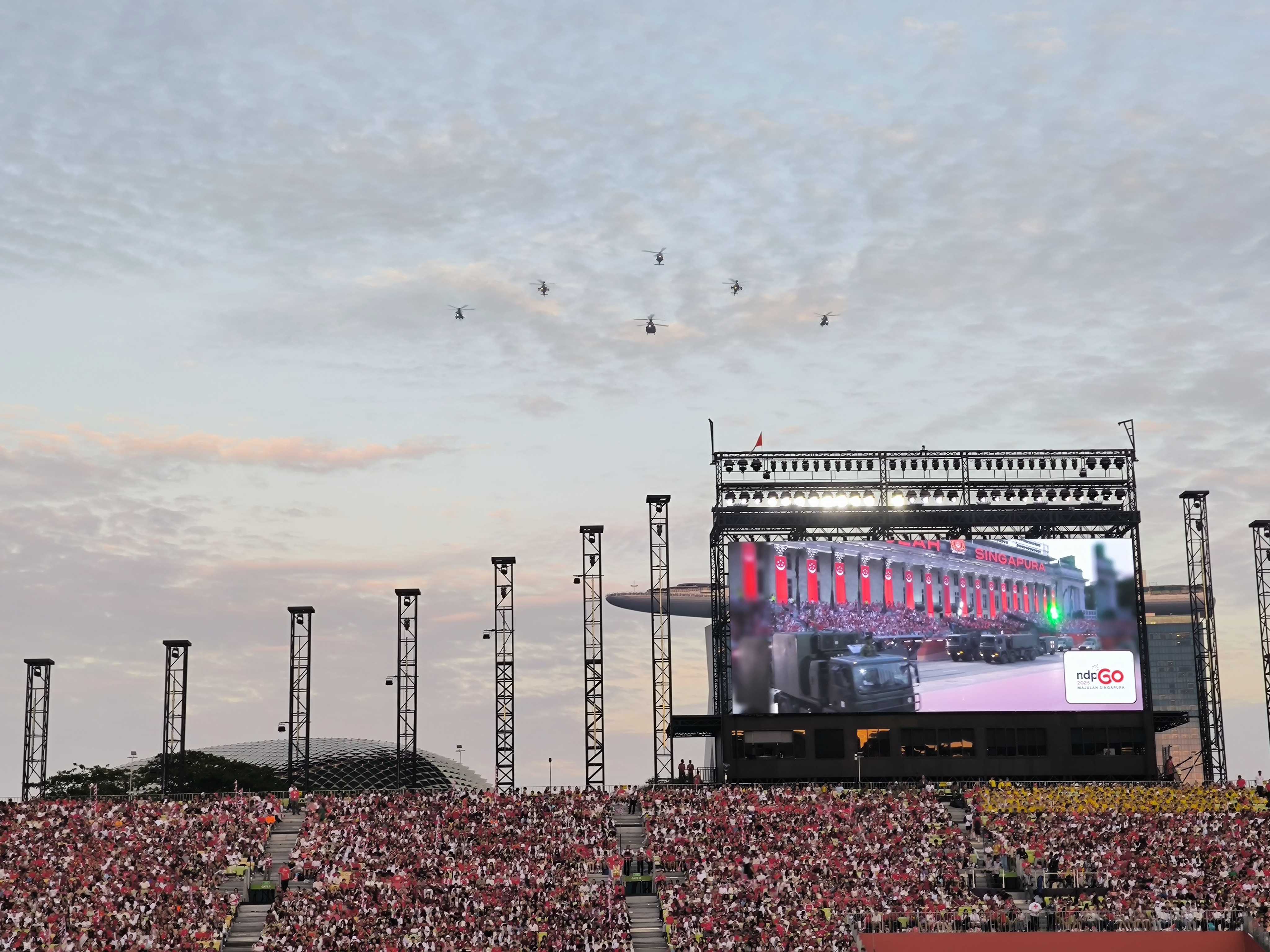 A packed stadium crowd watches a Singapore NDP parade, with helicopters flying overhead and a large screen showing military vehicles.