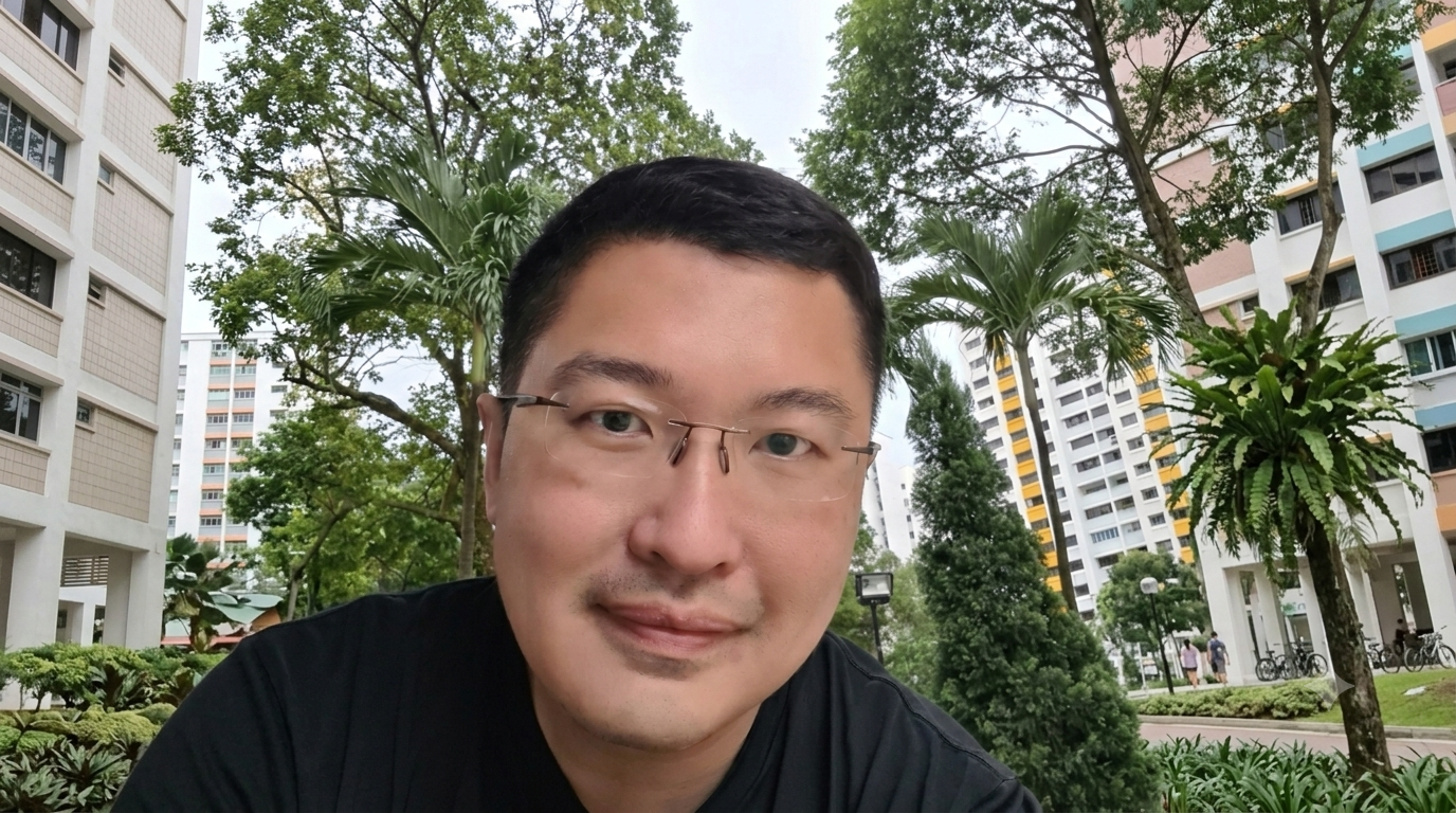 Middle-aged man with glasses and short hair outdoors, with trees and a tall building in the background.