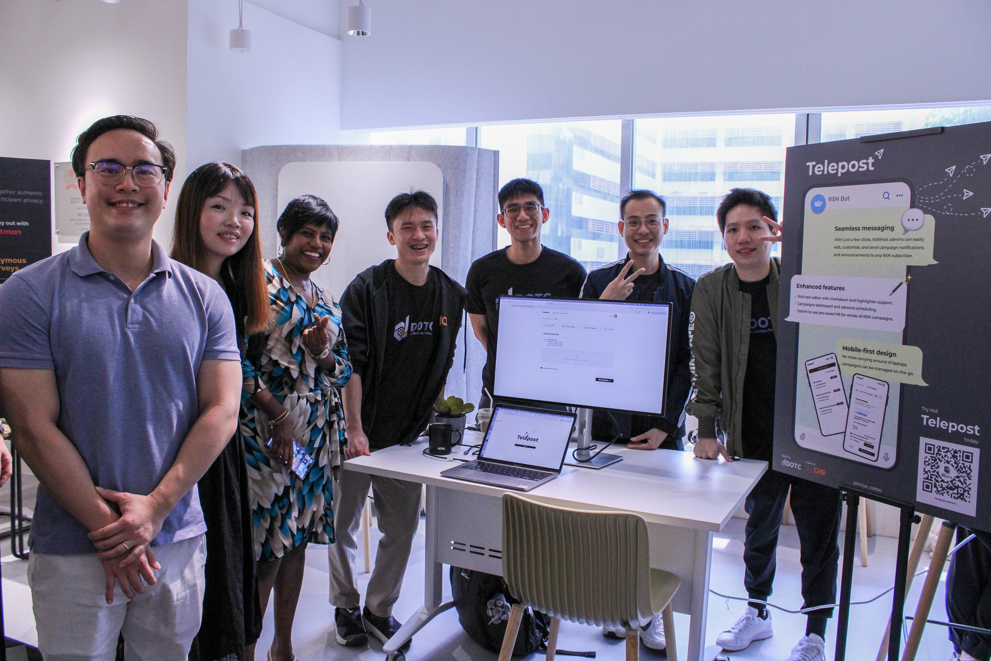 Seven people pose at a demo booth with a monitor and Telepost poster on an easel.
