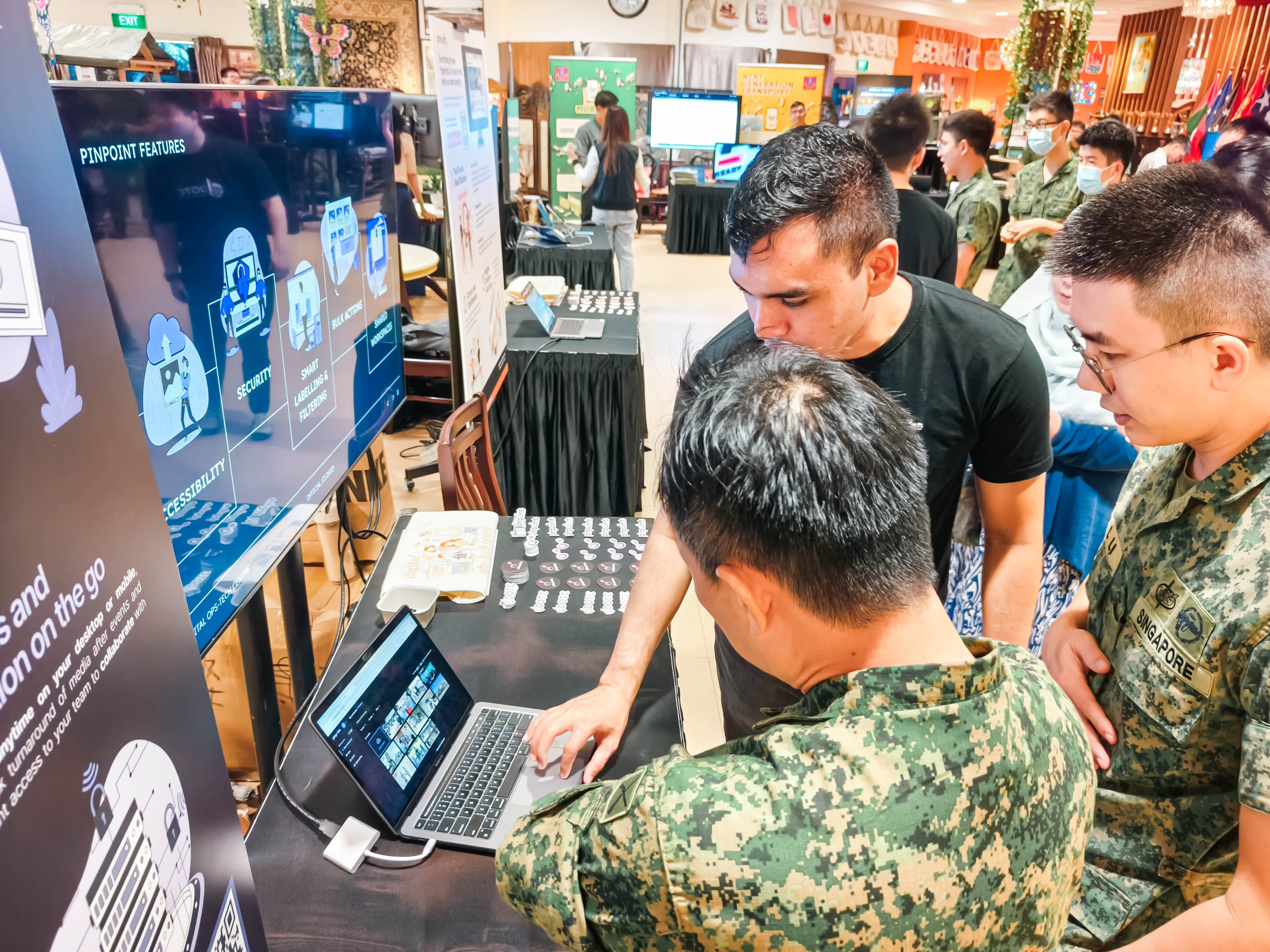 Three people, two in Singapore military uniforms, huddle around a laptop at a tech exhibition booth with a large display screen.