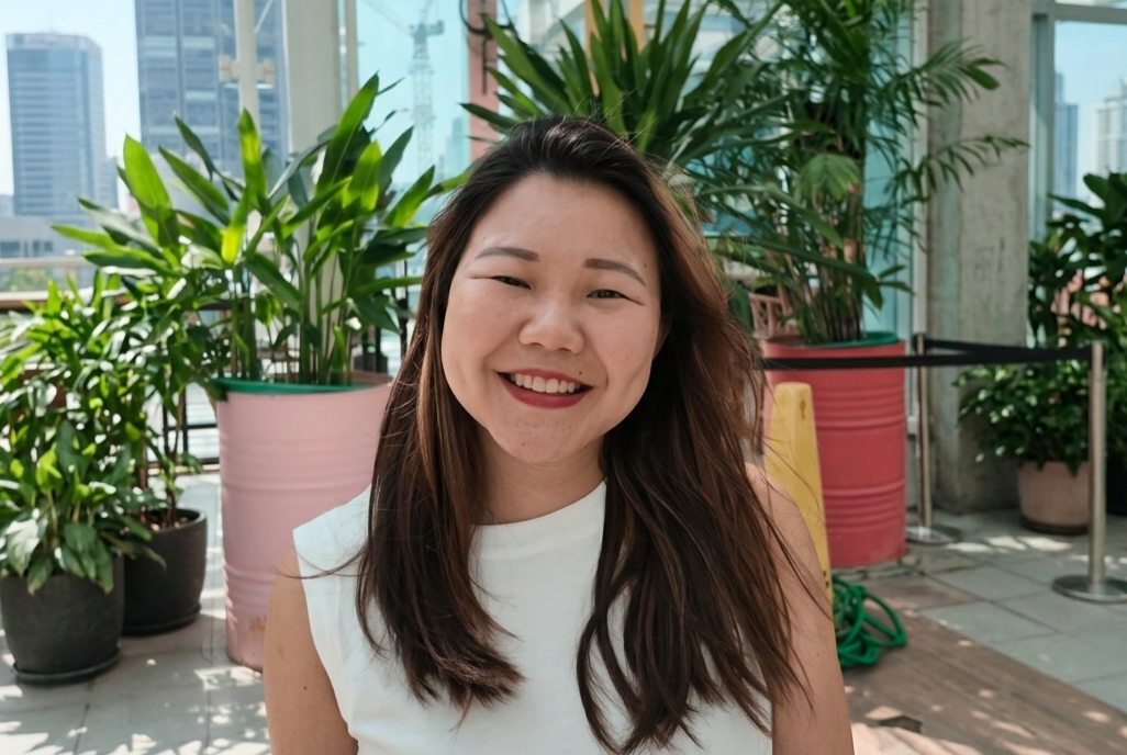 Smiling woman with long brown hair wearing a white top, seated outdoors on a rooftop with potted plants behind her.