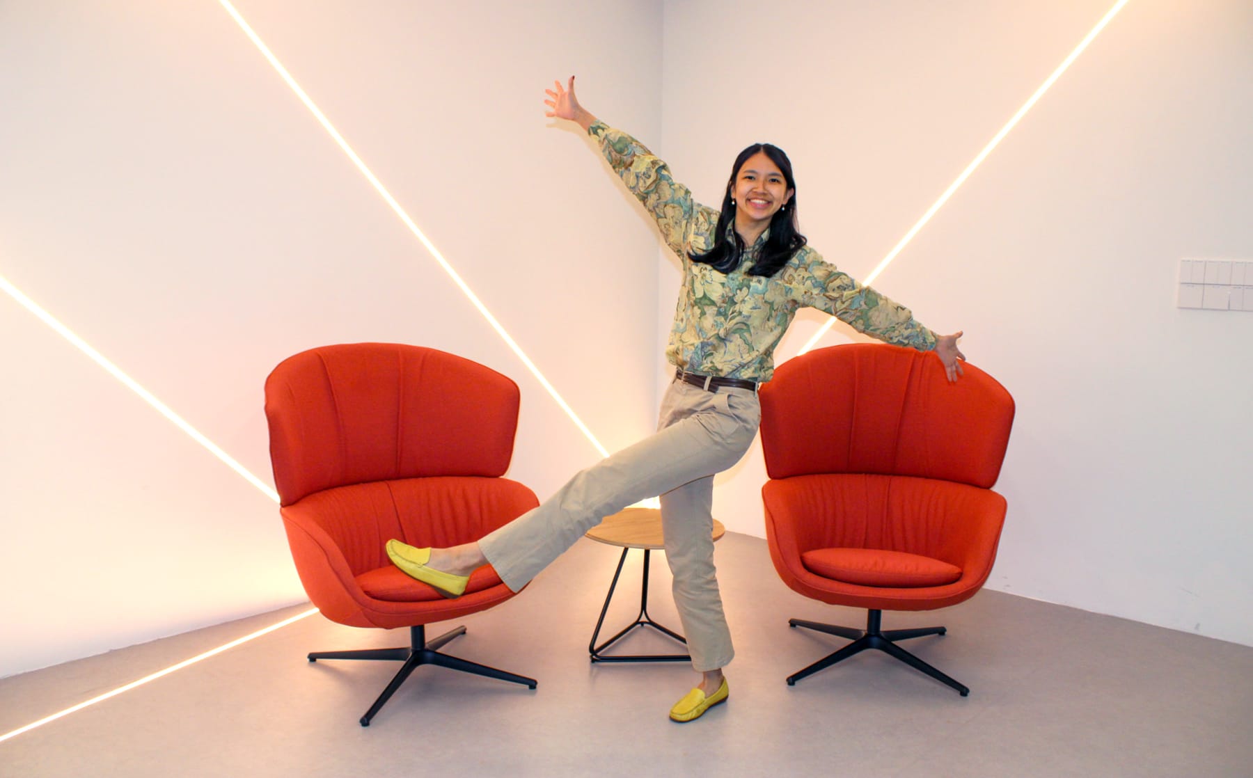 A smiling woman poses with one leg raised between two red armchairs, wearing a floral top and yellow loafers.