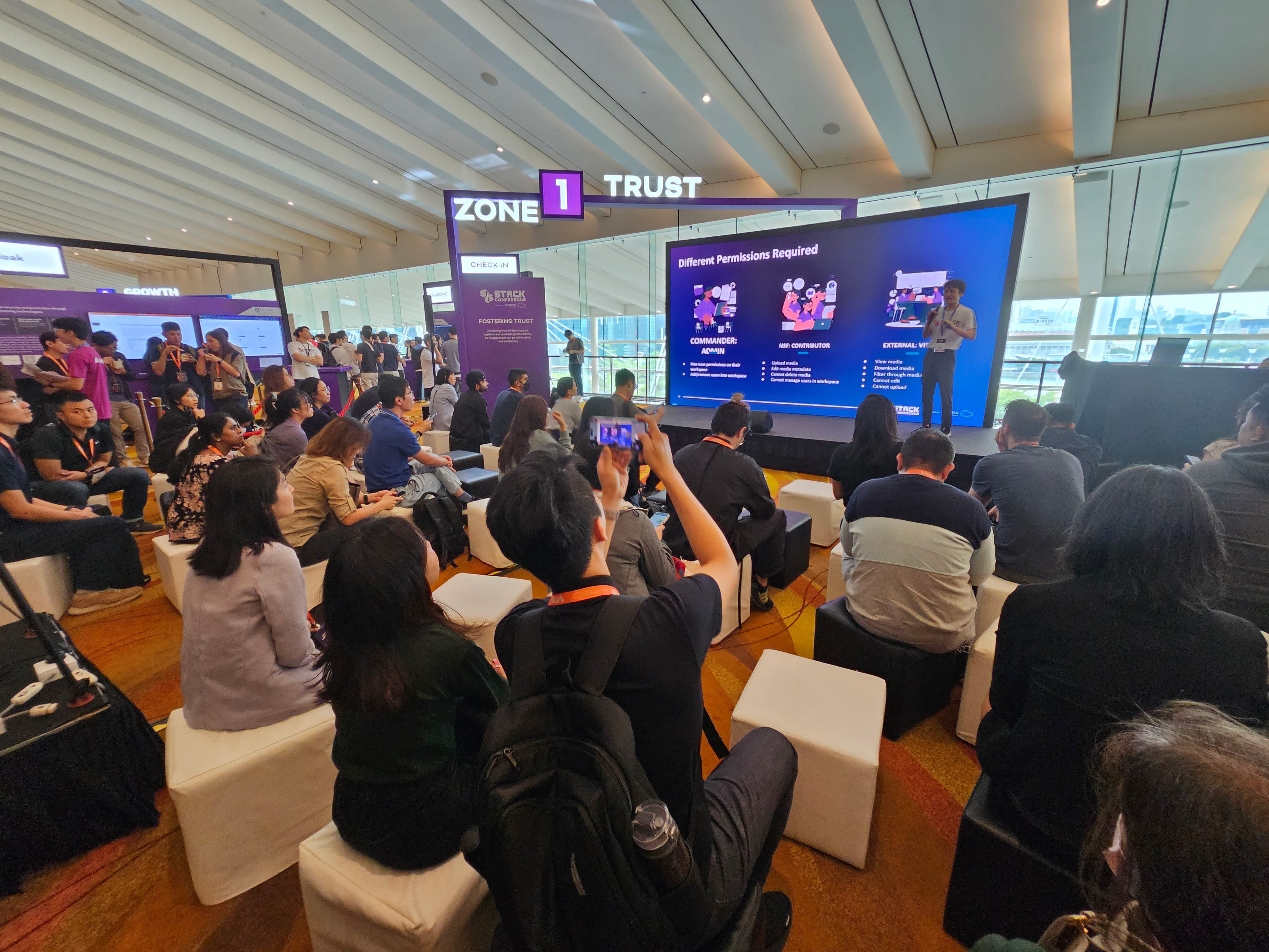 Audience seated on white stools at Zone 1 Trust conference, watching a speaker present a permissions slide on a large screen.