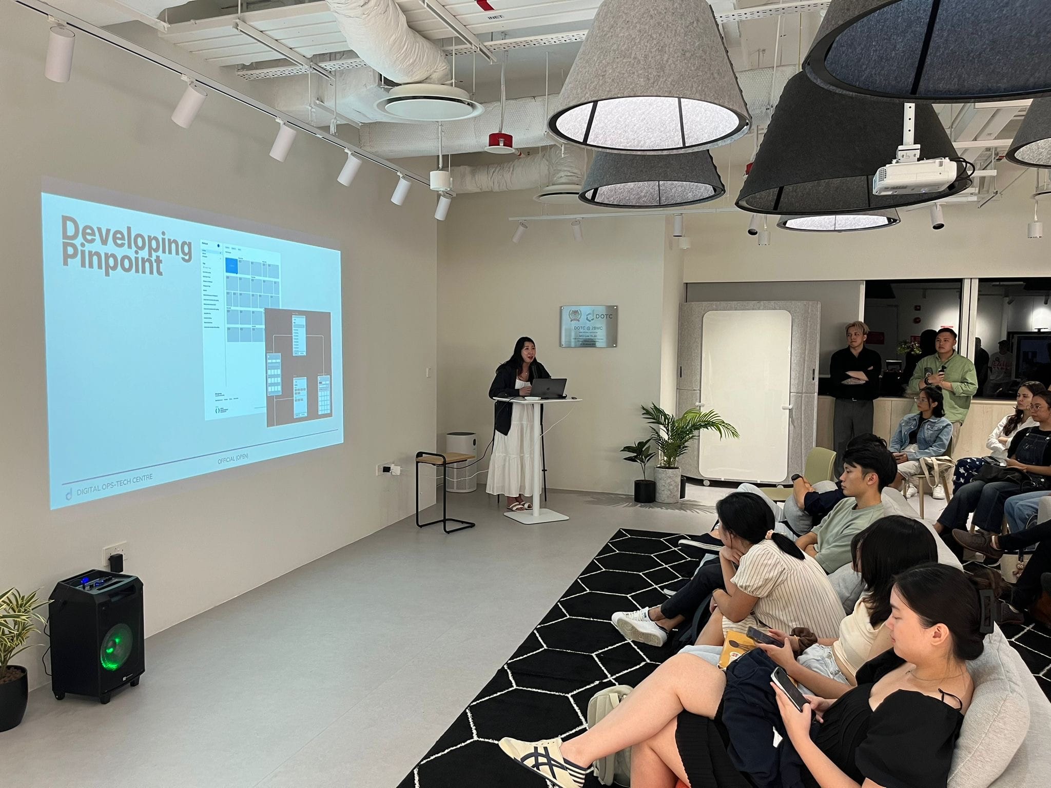 A woman presents at a podium before a projected slide reading "Developing Pinpoint" to a seated audience in a modern room.