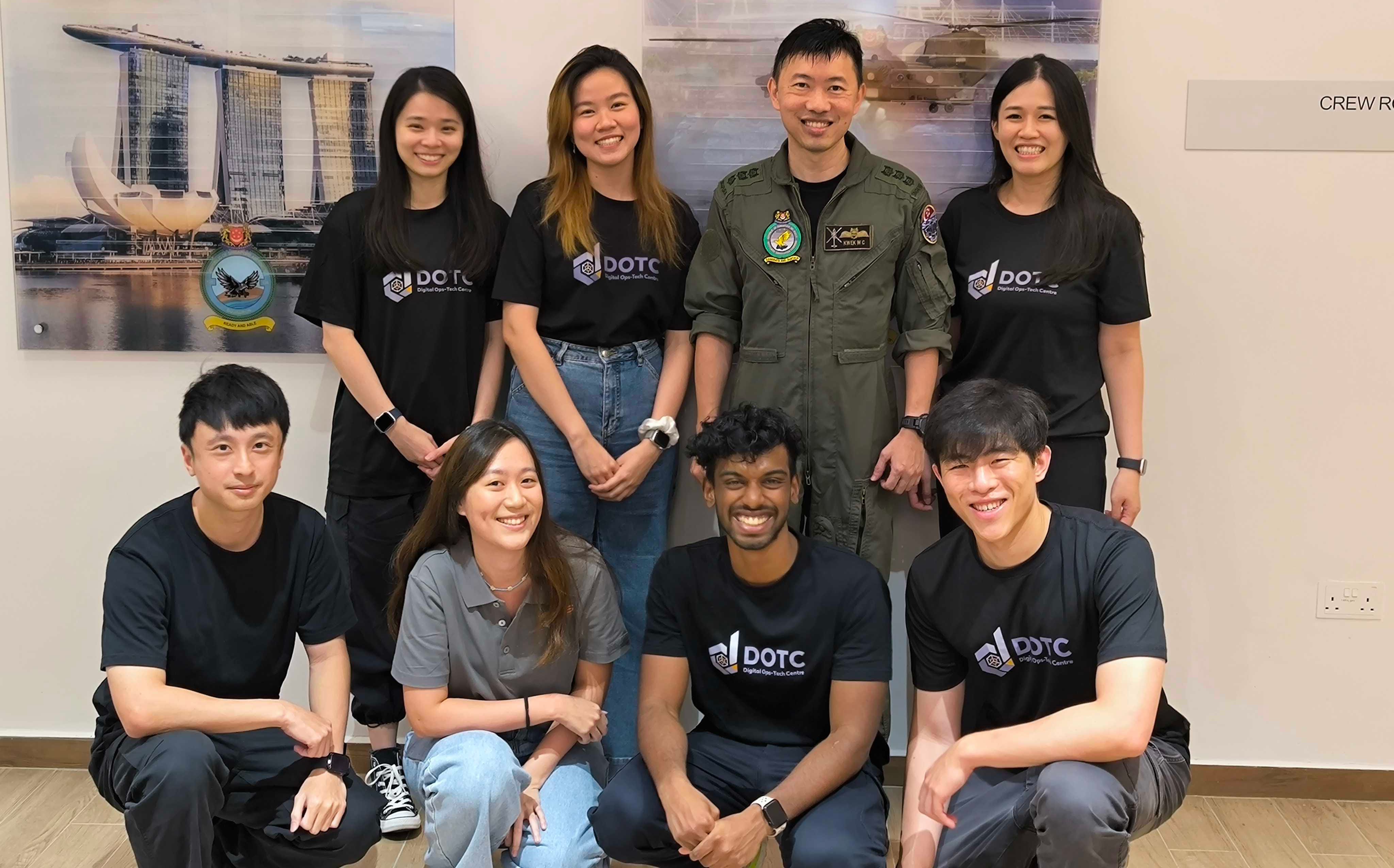 Eight people pose in two rows, most wearing DOTC shirts, with one person in a military flight suit.
