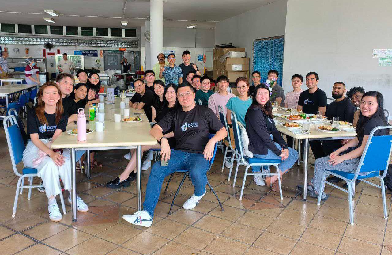 A large group of people in matching black DOTC shirts sit at tables with food in a cafeteria, smiling at the camera.