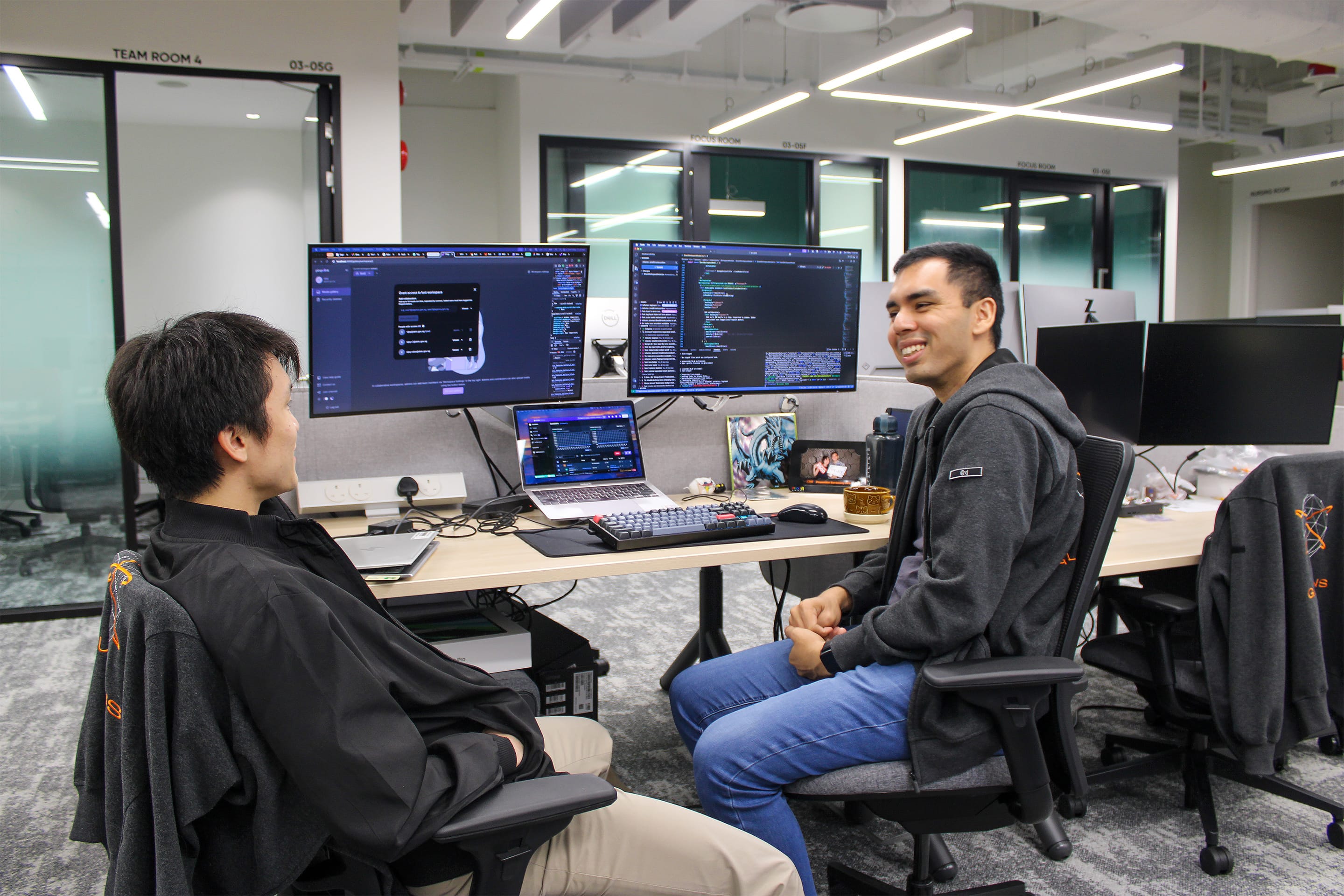 Two coworkers chat at desks in a modern office, with dual monitors and a laptop showing code.