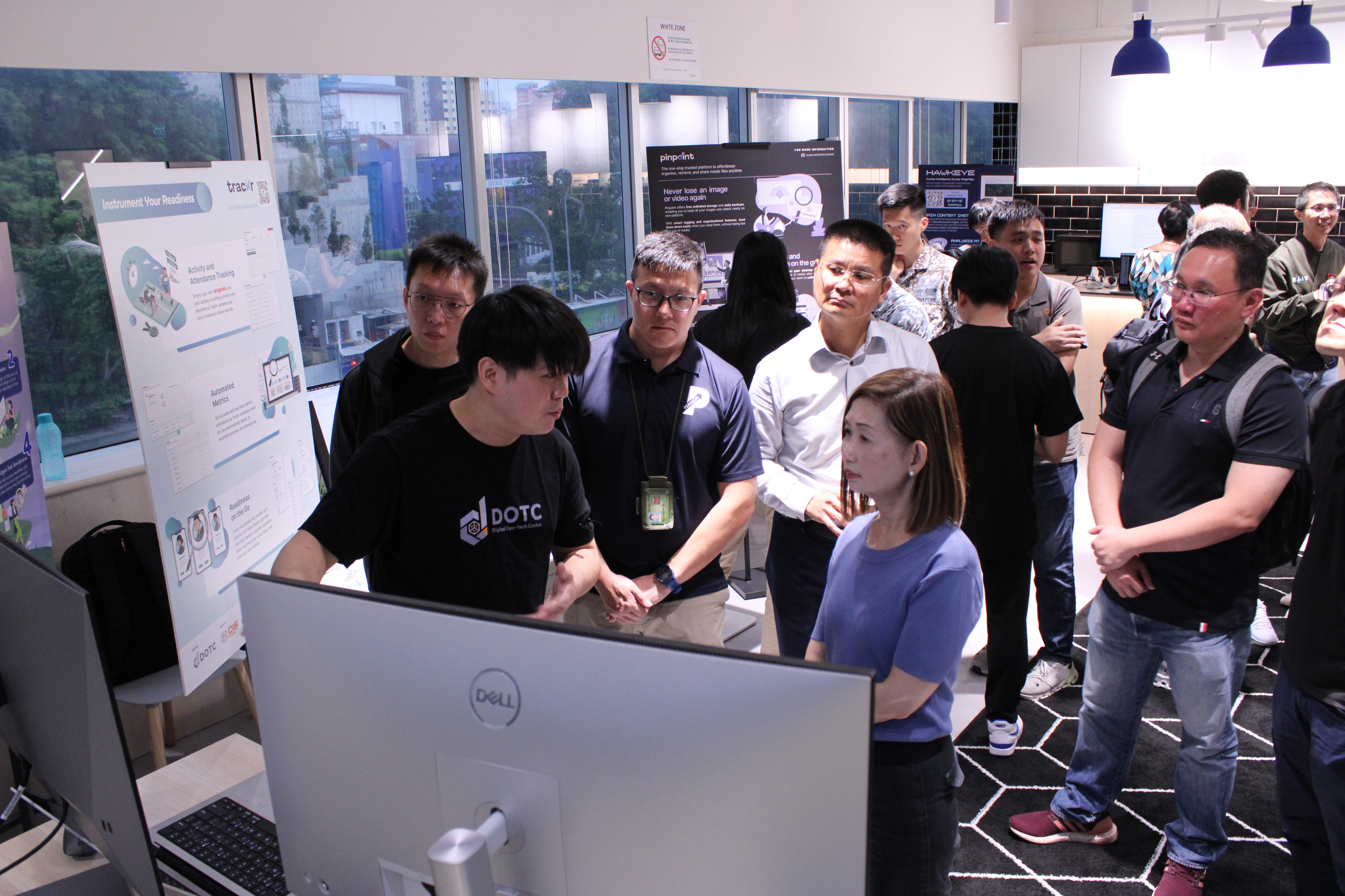 Group of people at a tech demo in an office, gathered around a Dell monitor and display posters.