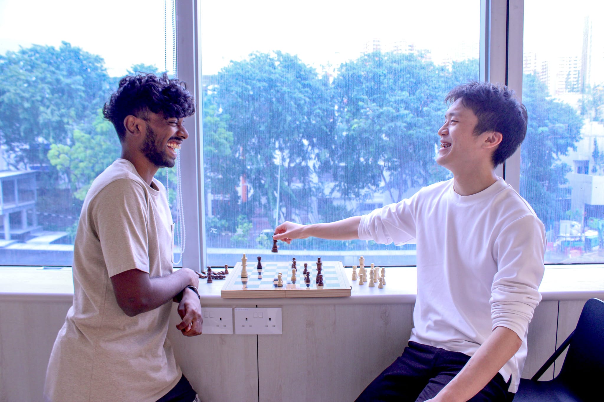 Two young men laugh while playing chess on a windowsill with trees visible outside.