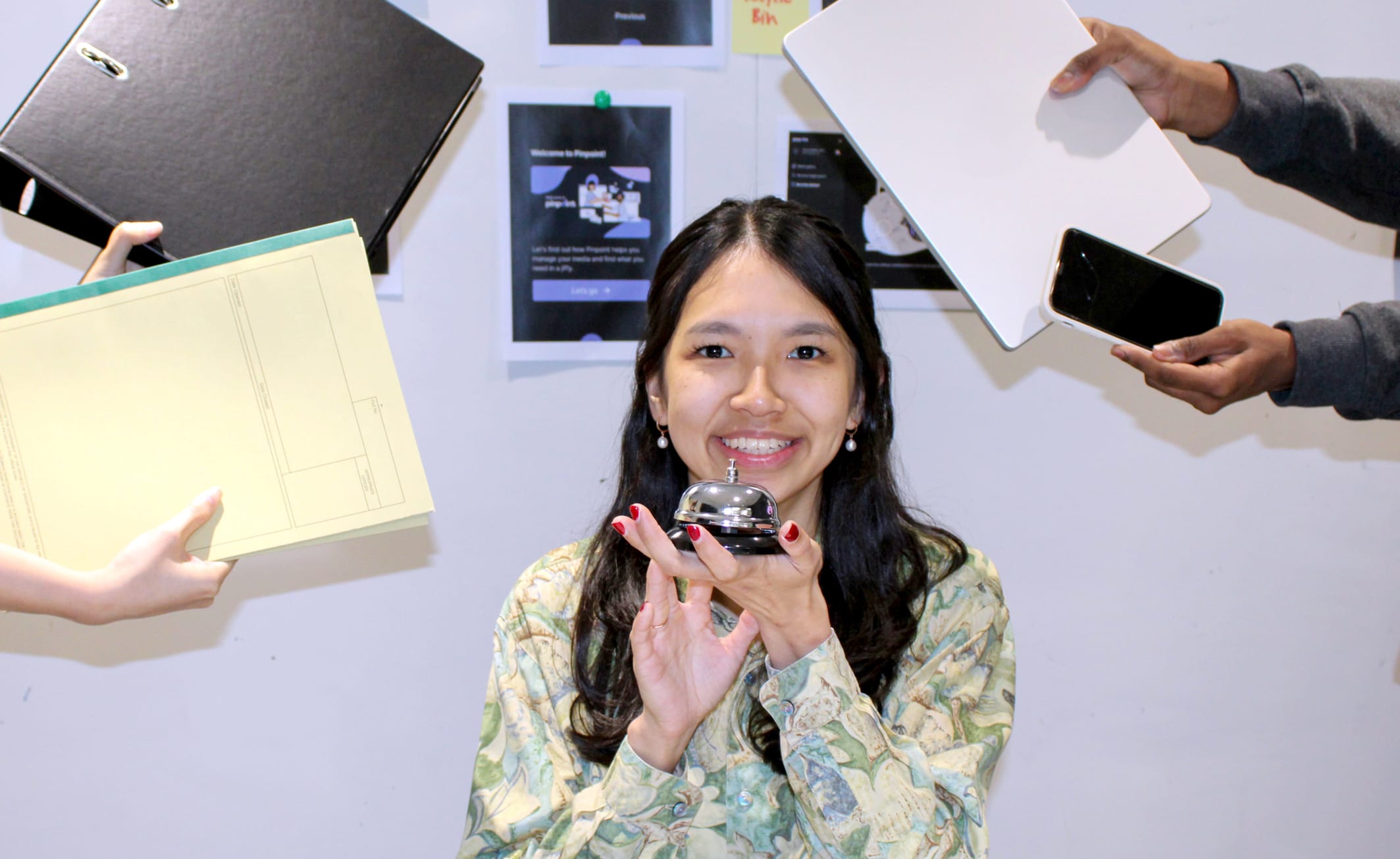 Smiling woman holding a desk bell, surrounded by hands offering folders, a tablet, and a phone near a pinboard.
