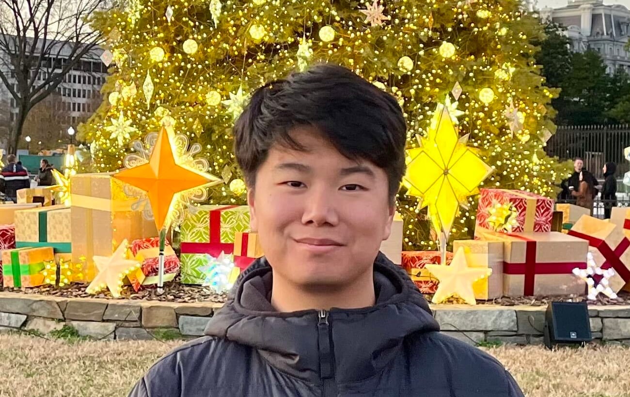Young man with short dark hair smiles at the camera, wearing a black jacket in front of a brightly lit Christmas tree and festive decorations.