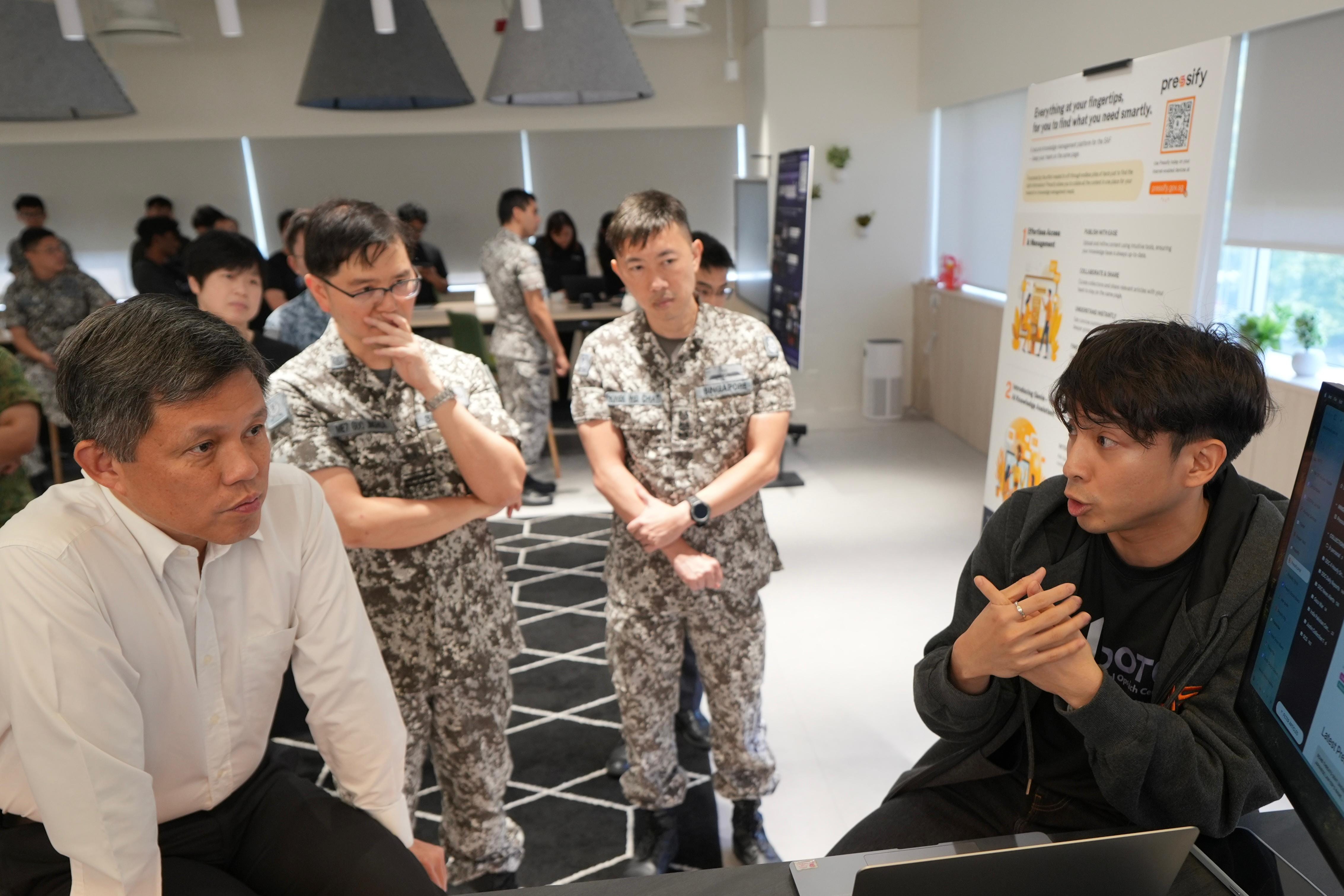 A man in a white shirt and two people in camouflage uniforms listen to a young man speaking near a monitor.