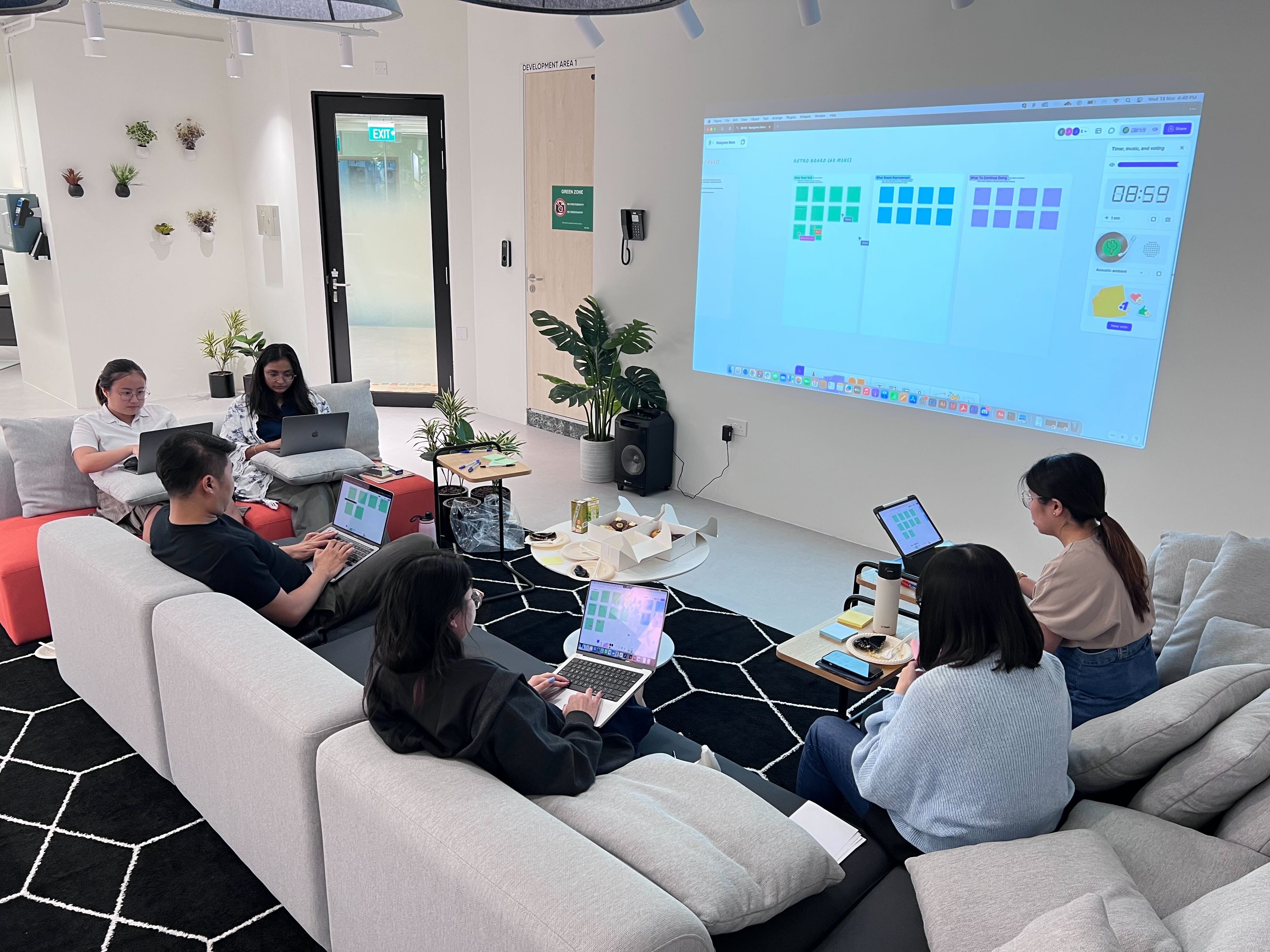 Five people sit on sofas with laptops in a modern lounge, facing a large projected screen showing a digital board.
