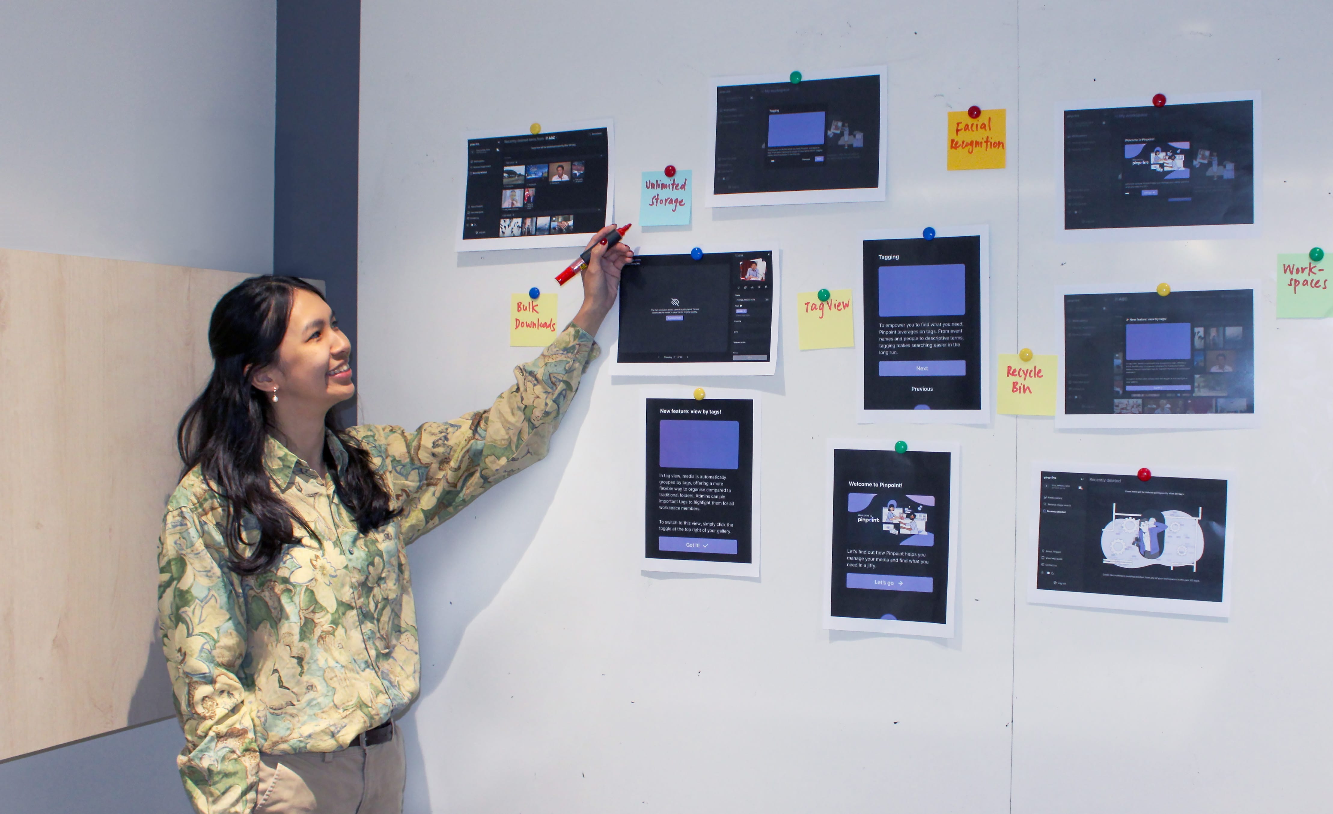 A smiling woman points with a red marker at printed screenshots and sticky notes pinned to a white wall.