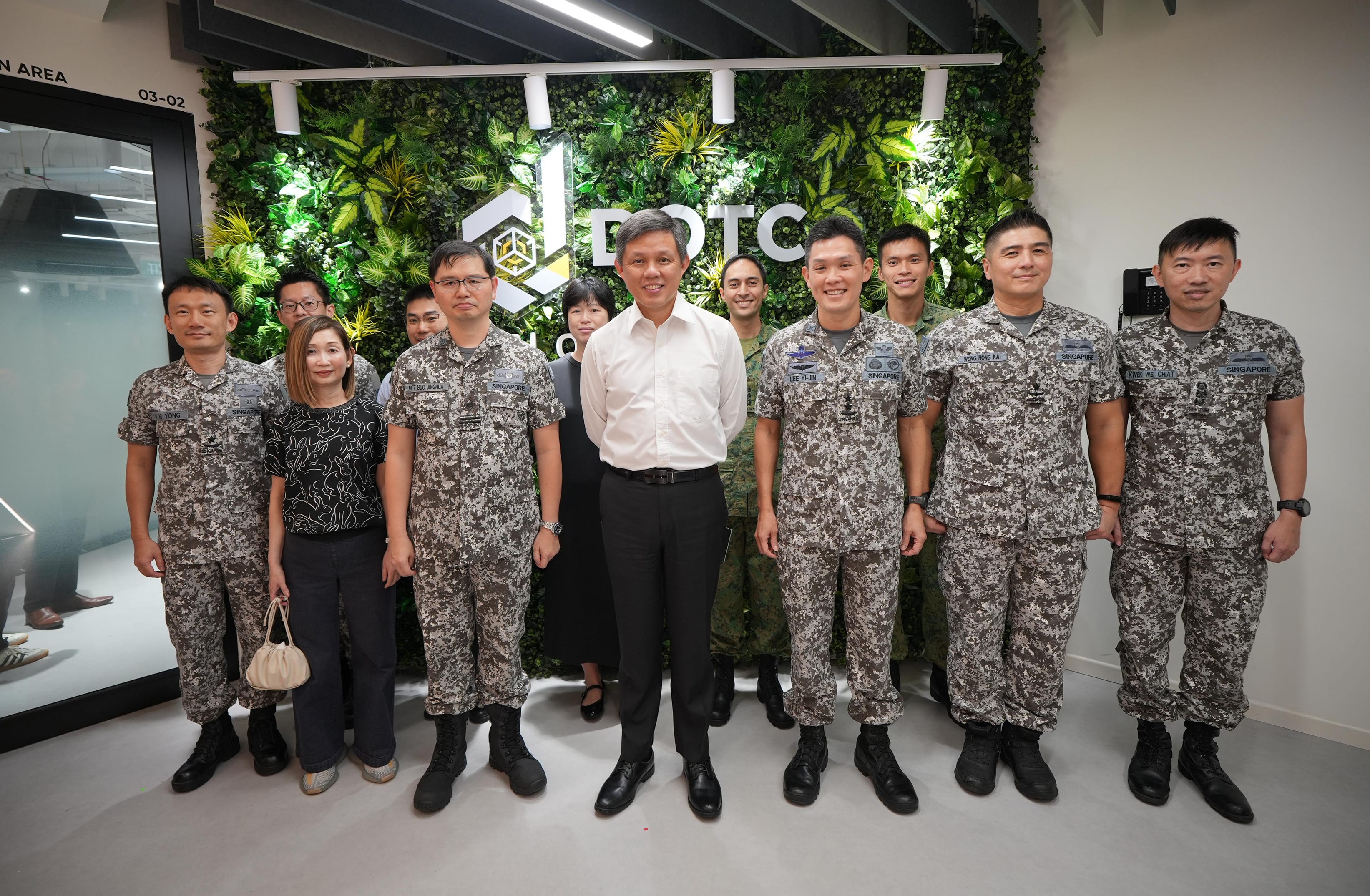 Group of uniformed soldiers and two civilians pose indoors before a green plant wall with DOTC sign