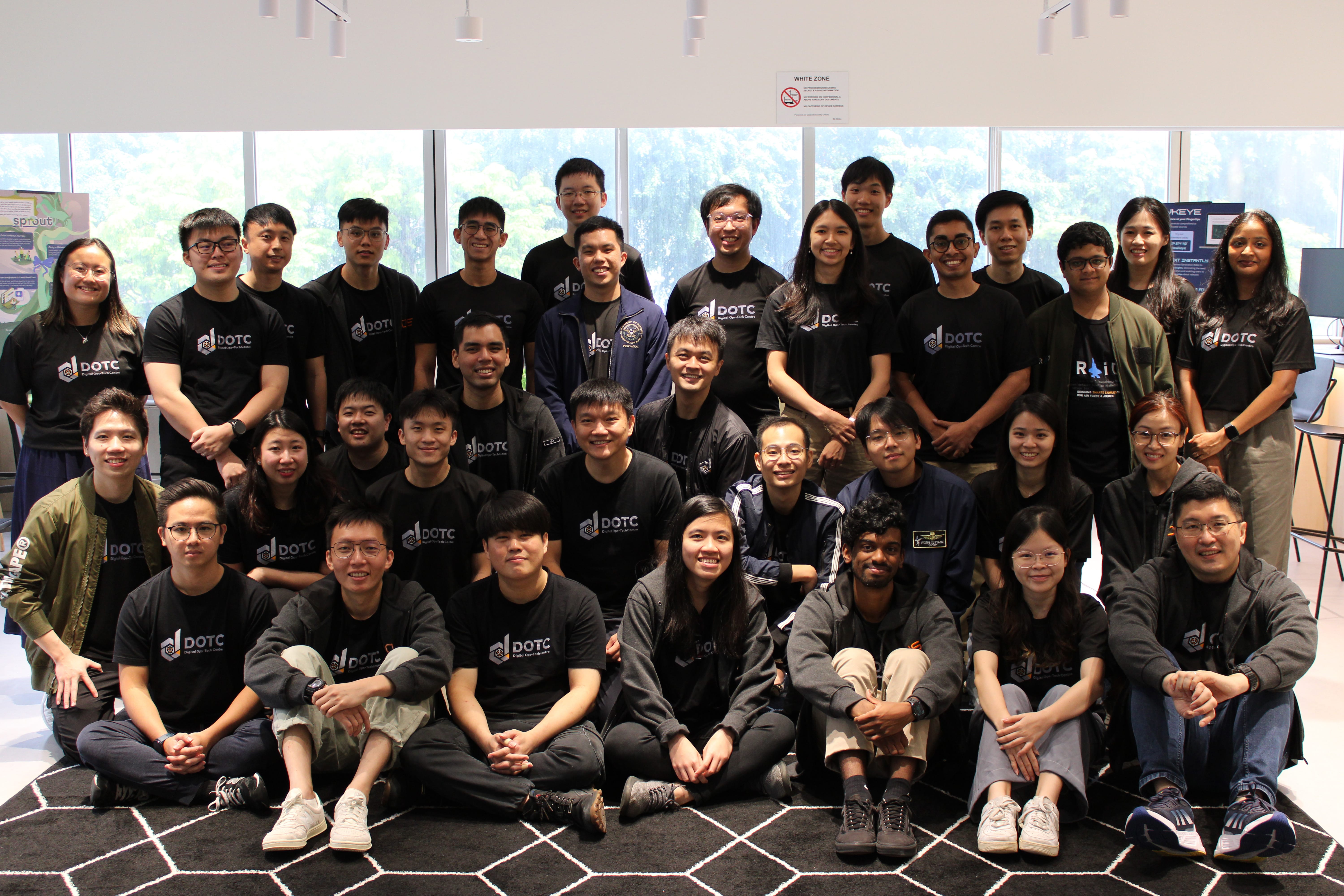 Group of about 25 people in black DOTC shirts posing indoors in front of large windows.