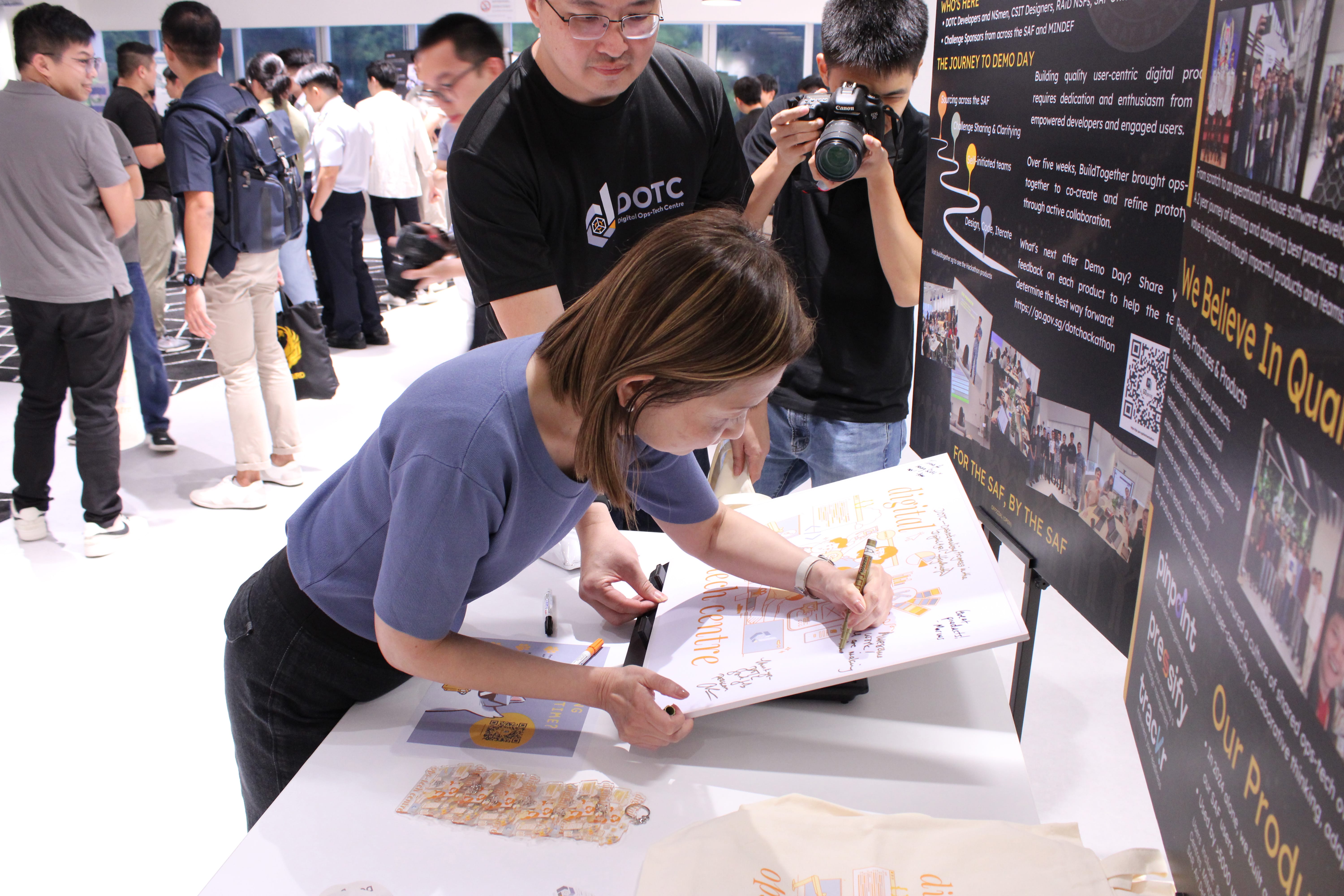 Woman leans over a table signing a poster as a man watches and another photographs her beside display boards.