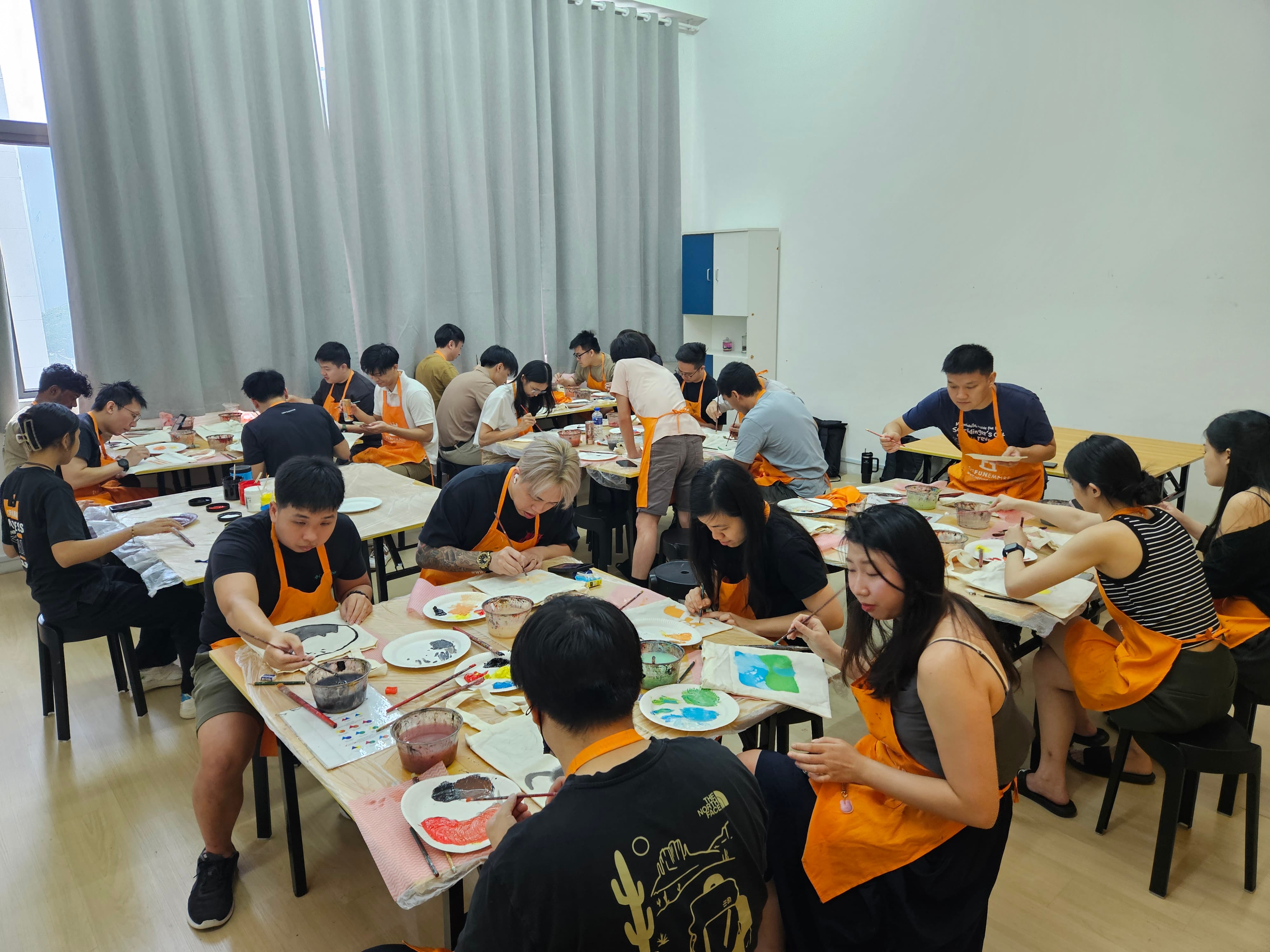 A group of people wearing orange aprons sit at tables in a bright room, painting on canvas during an art workshop.