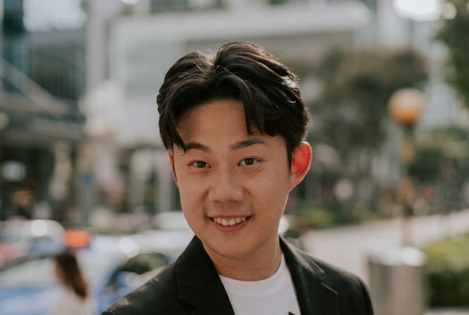 Young man with black hair smiling, wearing a black blazer over a white shirt, with a blurred street background.