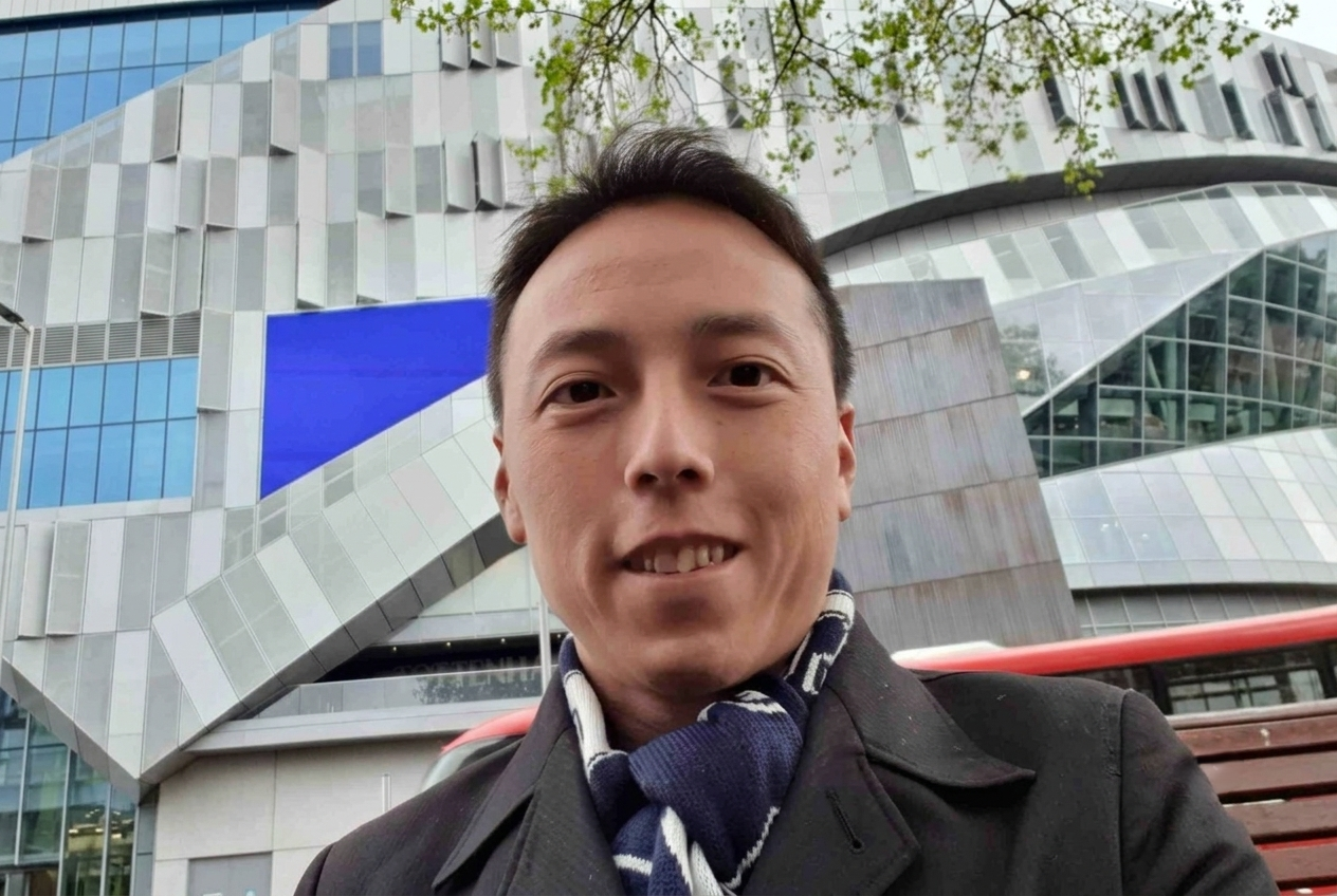 A close-up selfie of a smiling East Asian man wearing a dark coat and a blue and white patterned scarf, with a large, modern grey and blue building in the background.