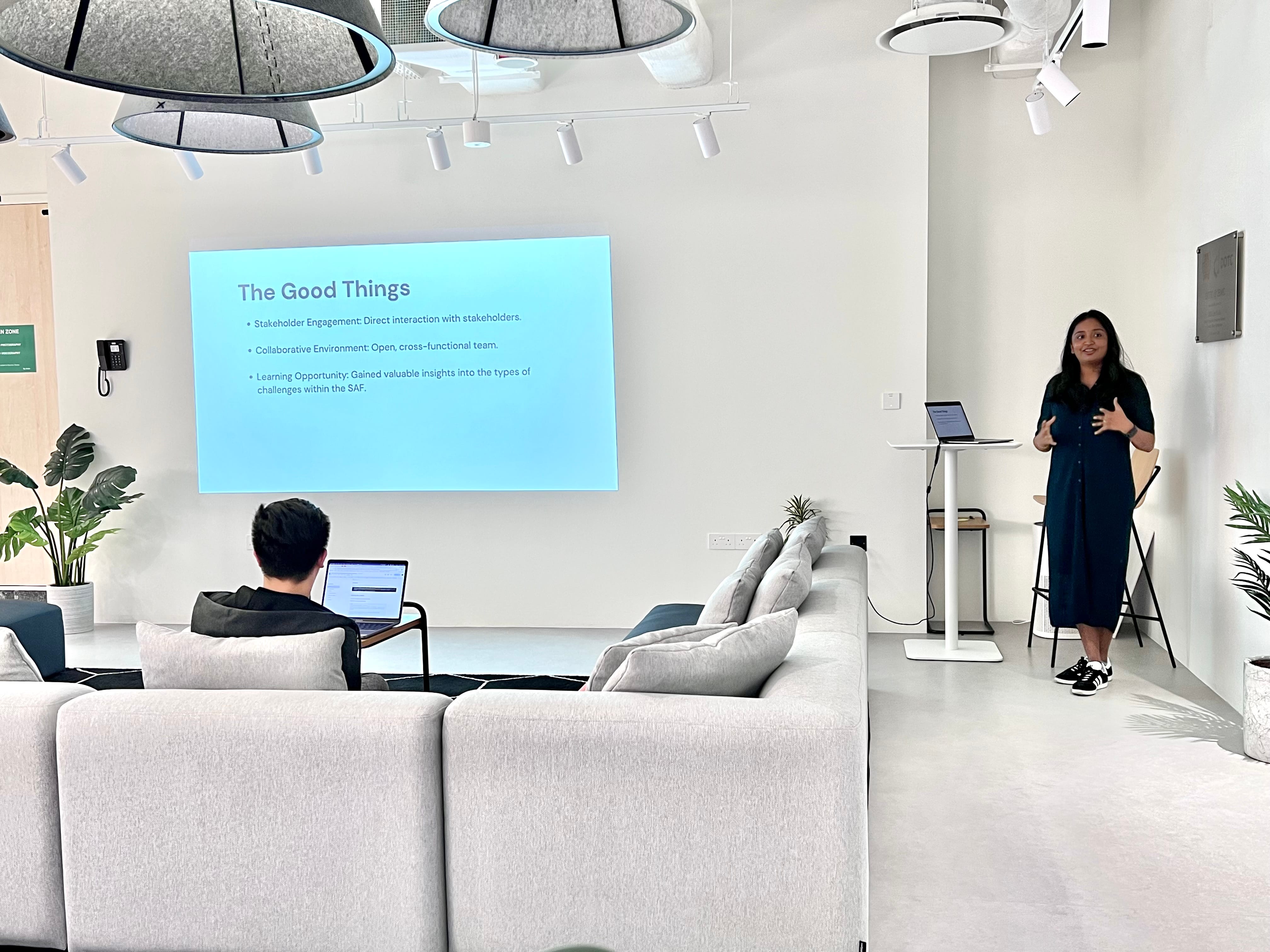 Woman presenting a slide titled "The Good Things" in a modern room, with a person seated on a sofa using a laptop.
