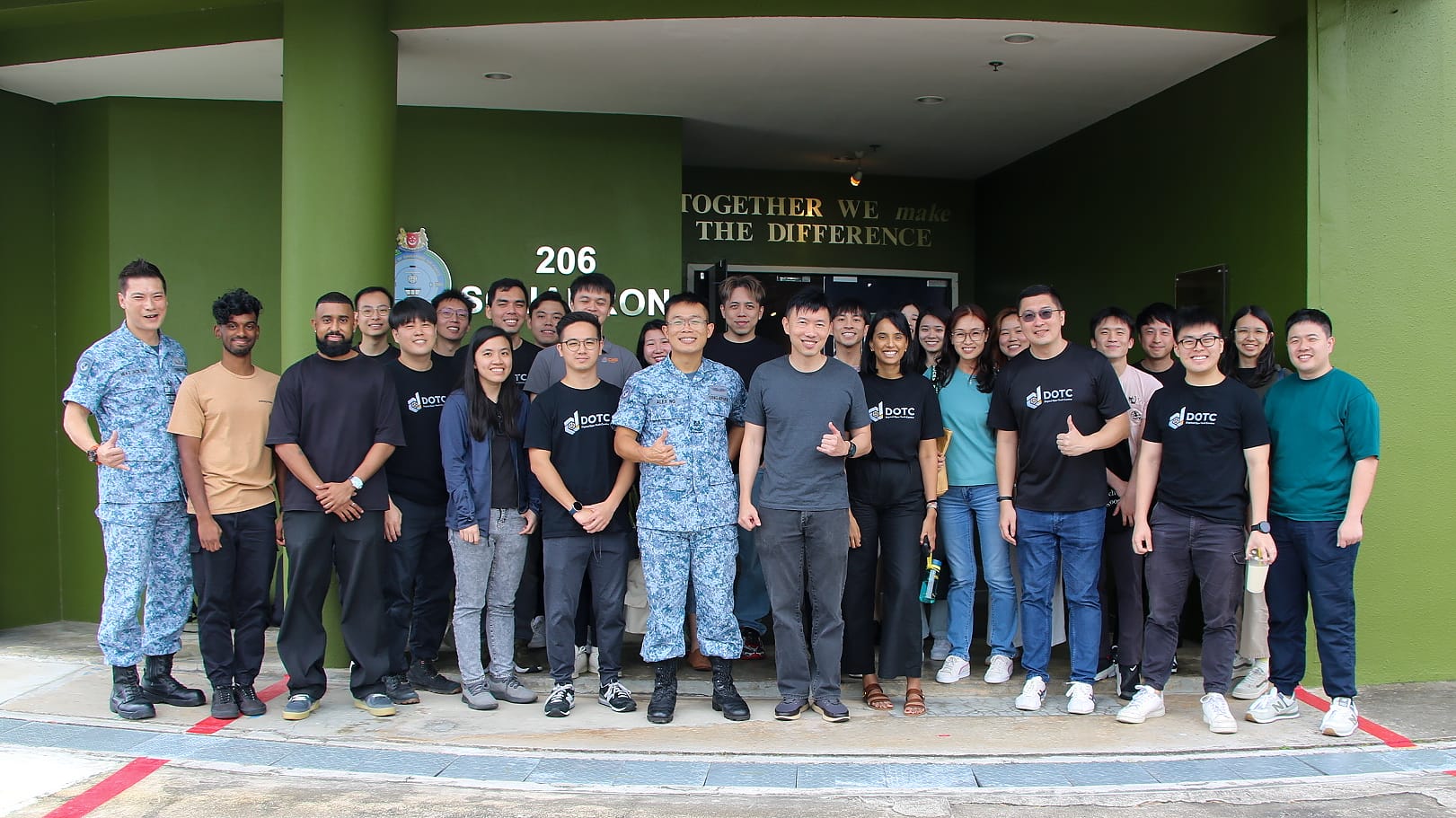 A large group of people, some in DOTC shirts and military uniforms, posing outside a green building labeled 206 Squadron.
