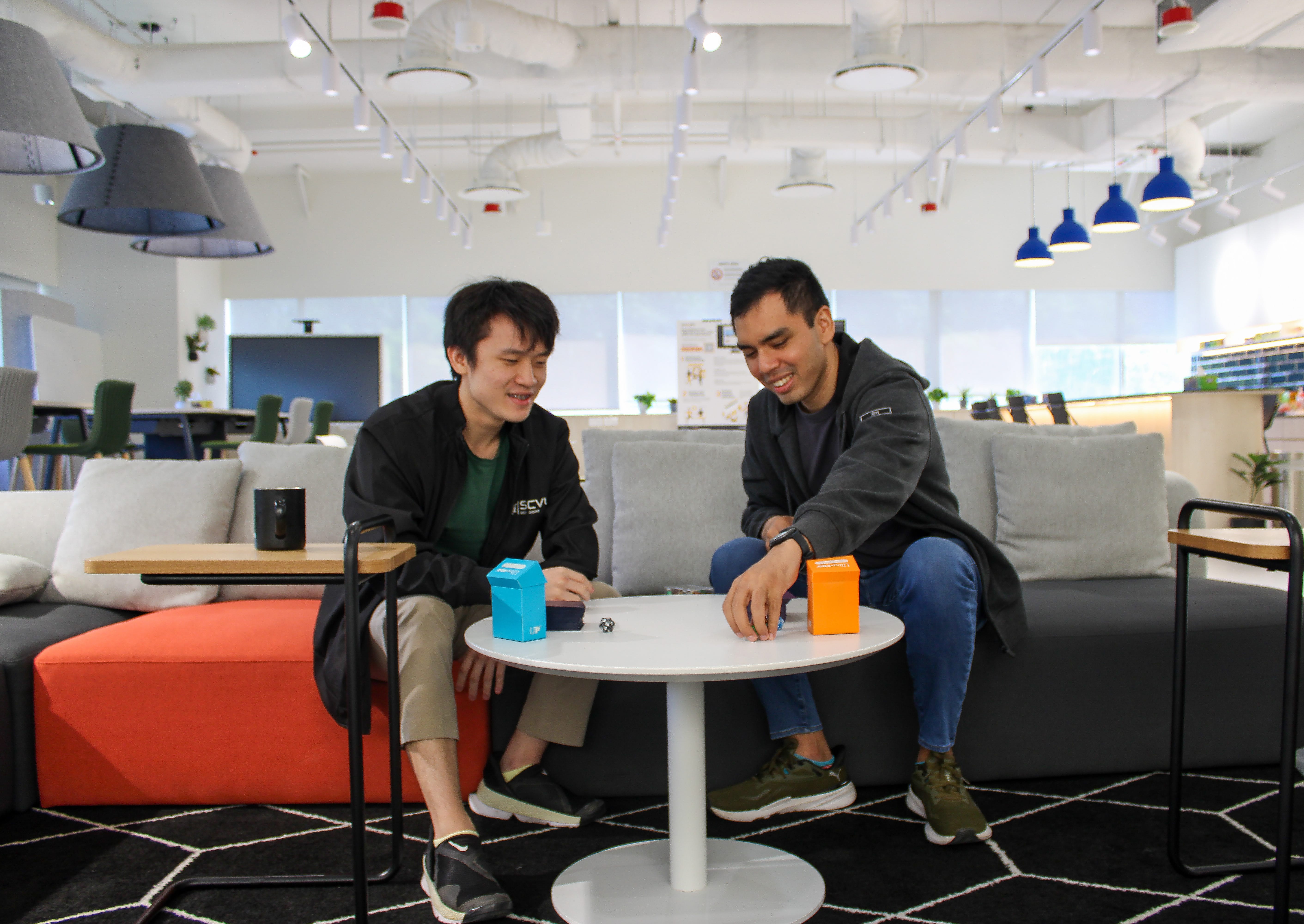 Two men sit on a couch in a bright office lounge, playing a tabletop game with blue and orange boxes.