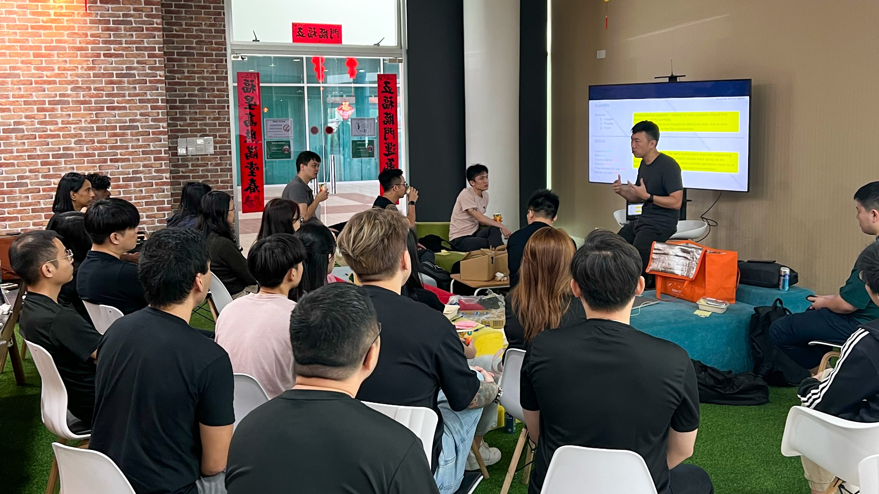 A man presents to a seated group in a room with brick walls, Chinese banners, and a screen showing highlighted text.