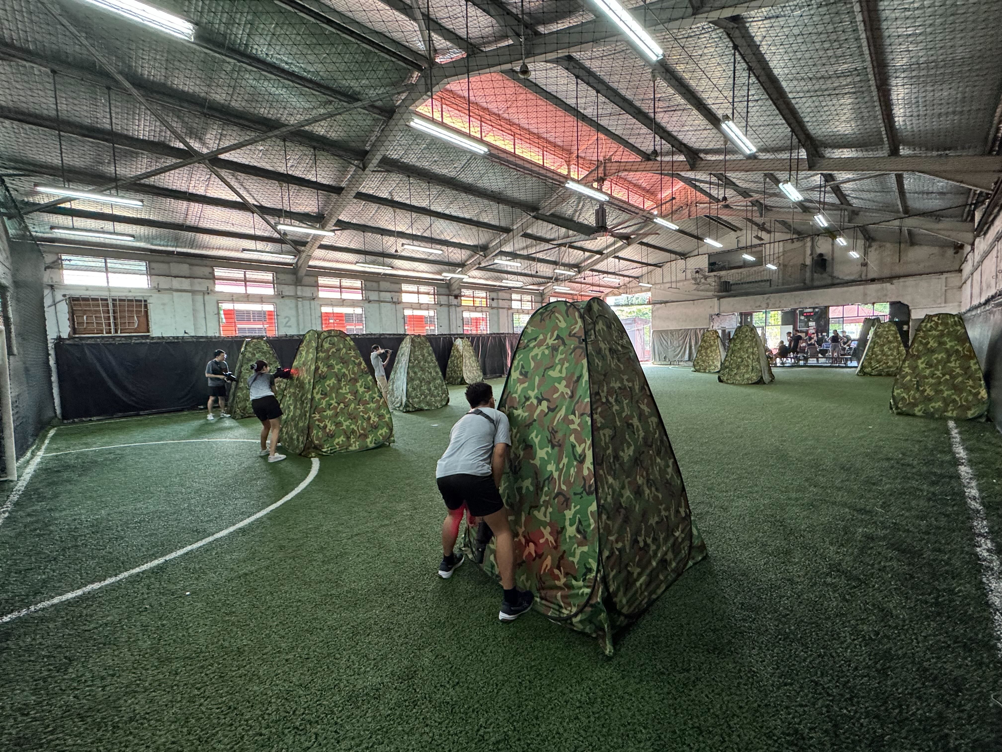 Indoor arena with green turf and camouflage pop-up tents used as cover for a laser tag or nerf battle game.