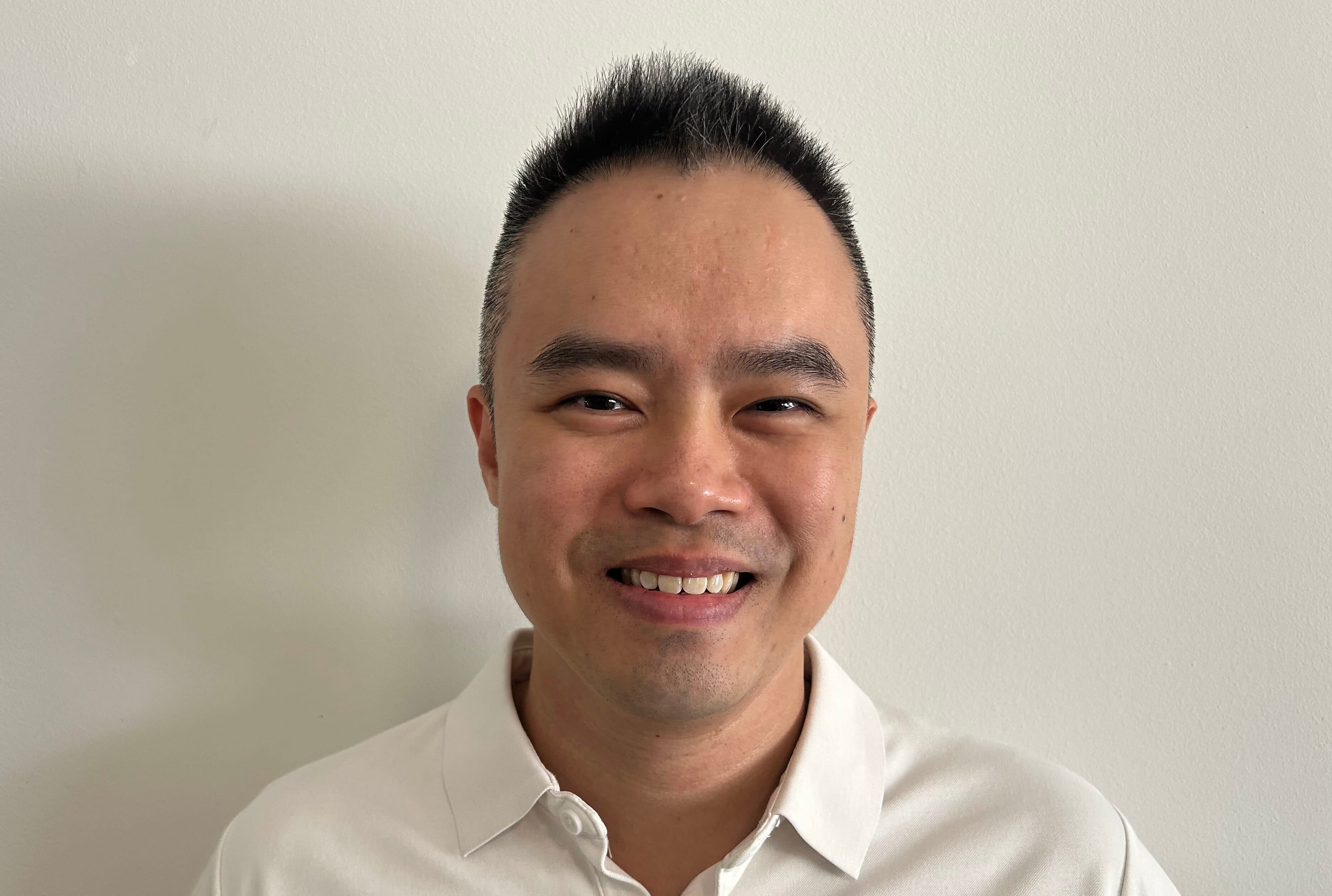 Man with short dark hair smiles at the camera, wearing a white collared shirt against a plain white wall.