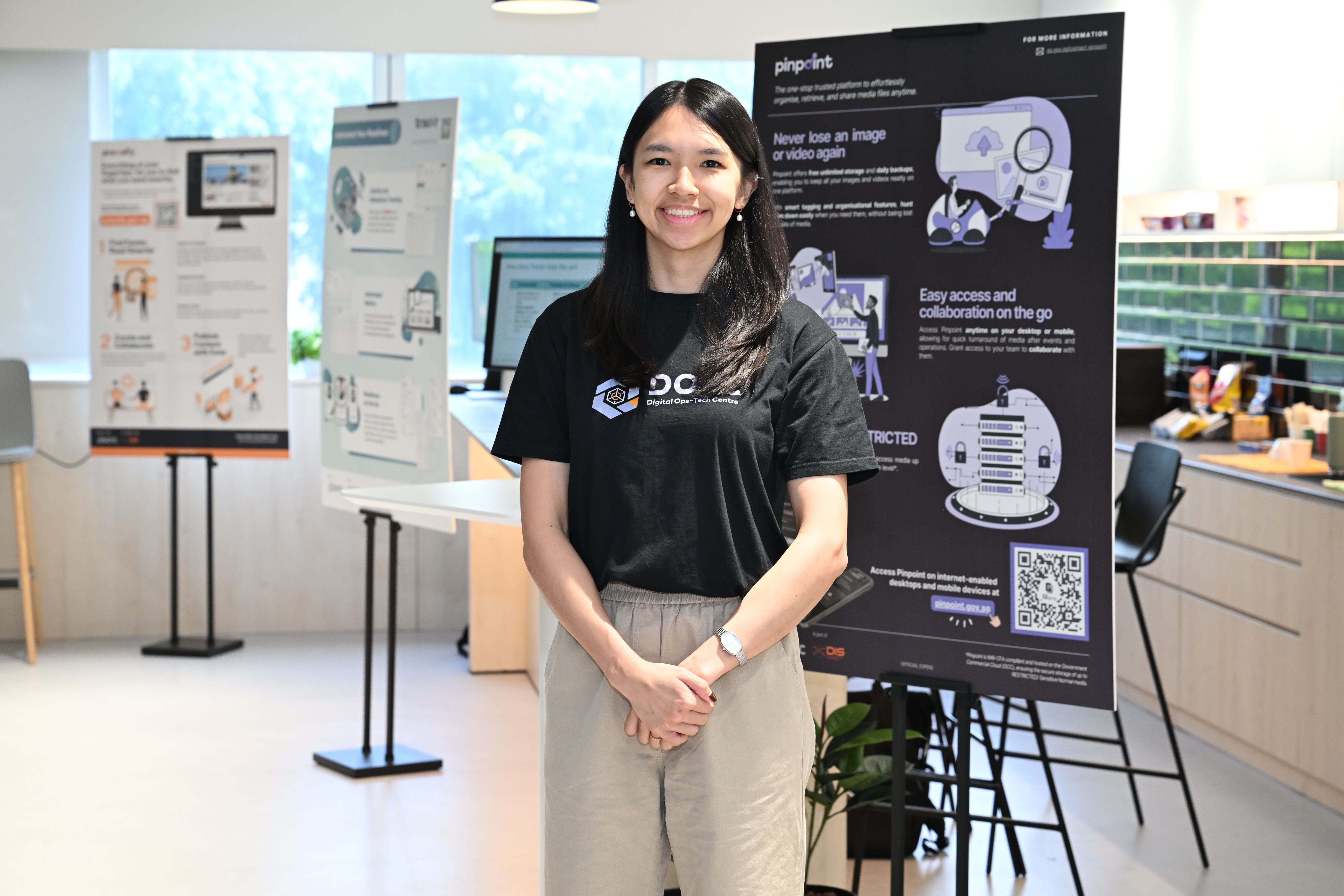 Young woman in a black Digital Ops-Tech Centre t-shirt smiling in front of a Pinpoint product display board.
