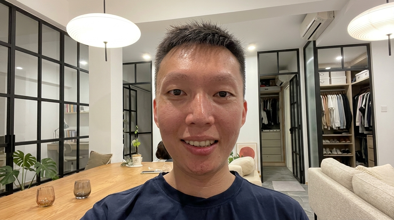 Young Asian man with short black hair smiling, wearing a dark shirt, indoors with modern interior behind him.