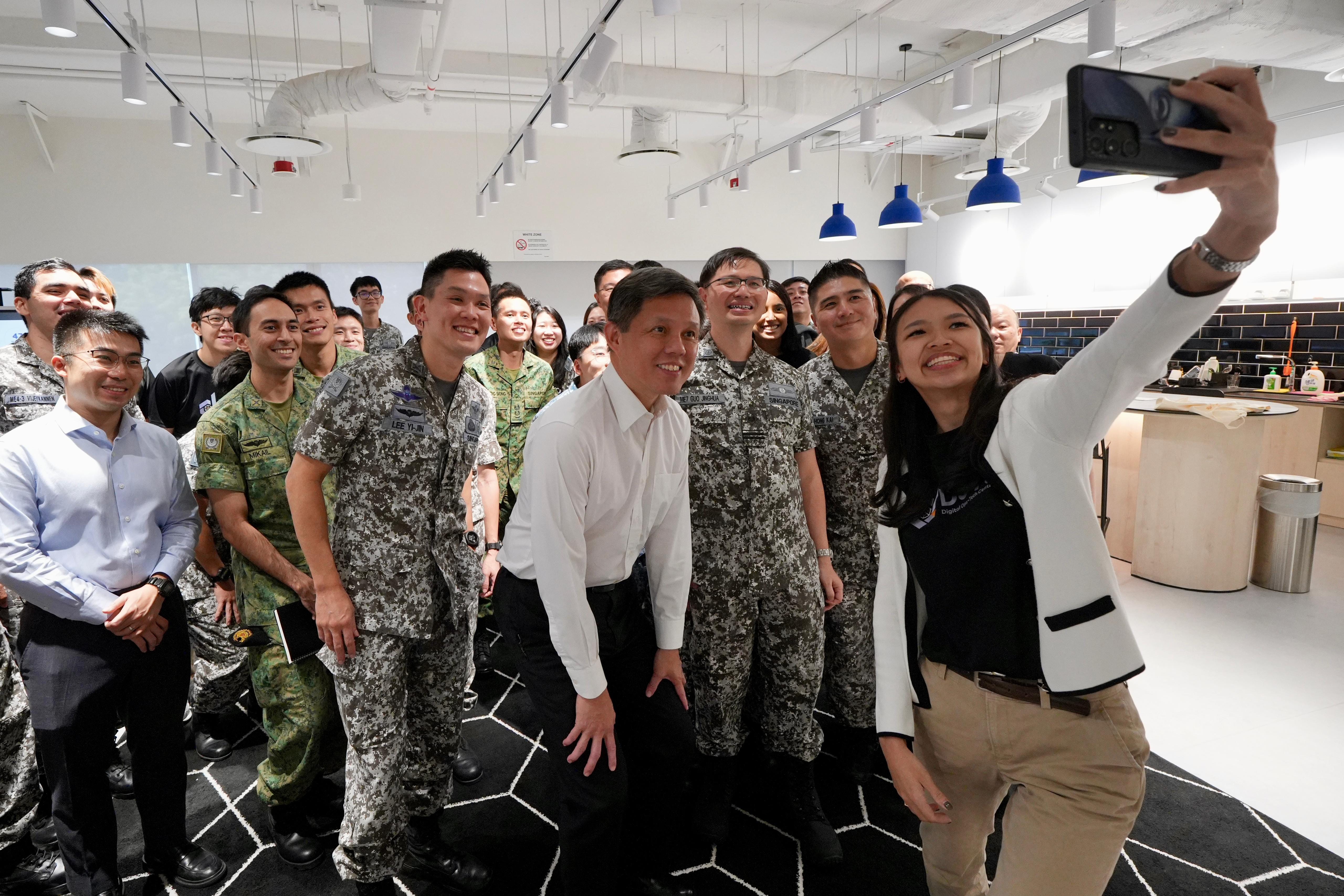 A woman takes a selfie with a large group including military personnel in camouflage and civilians indoors.