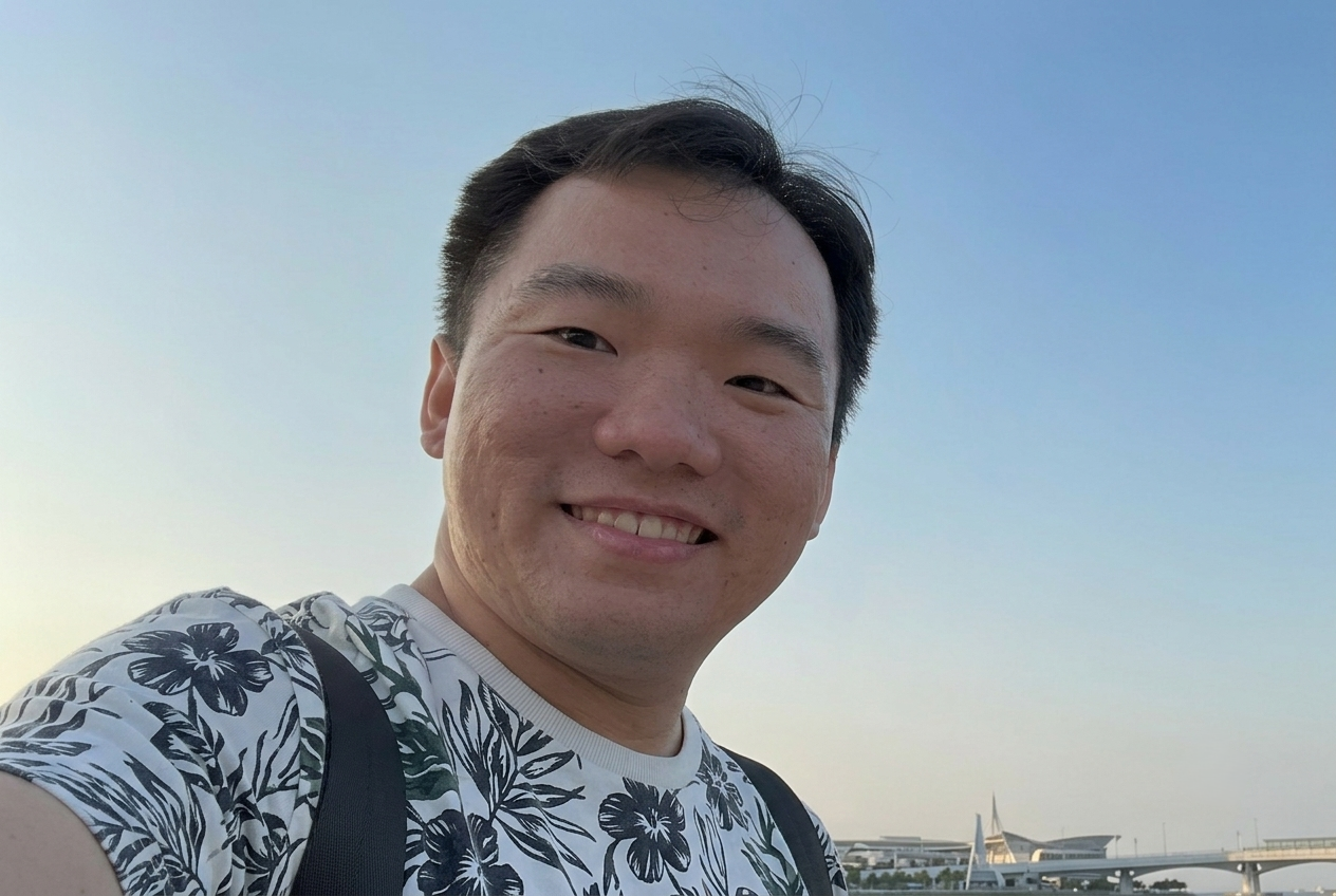 A close-up selfie of a smiling East Asian man with short dark hair, wearing a white t-shirt with a black floral pattern and backpack straps, set against a clear blue sky.