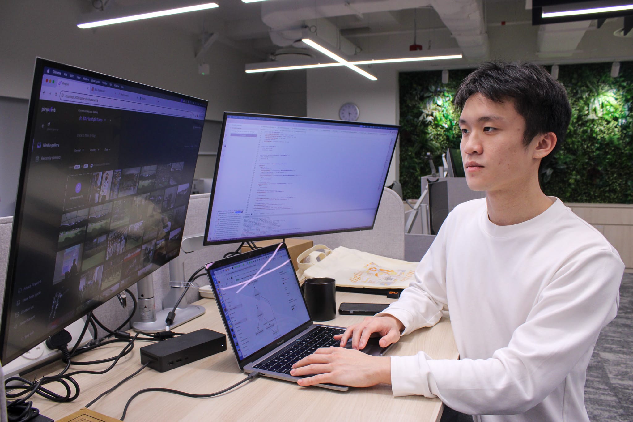 Young man in a white sweater working at a desk with a laptop and two external monitors showing code and media.