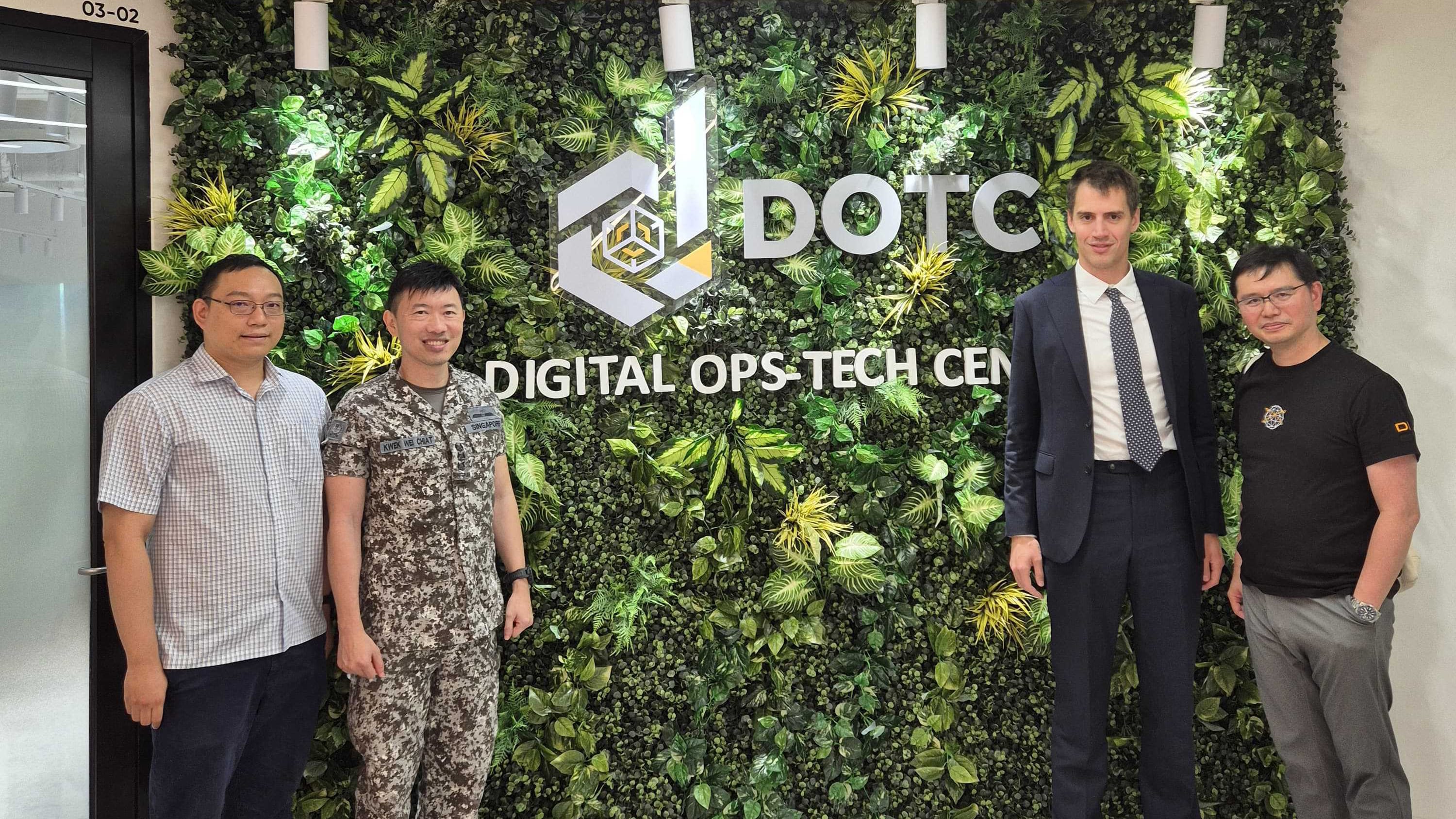 Four men stand before a green plant wall with the DOTC Digital Ops-Tech Centre sign and logo.