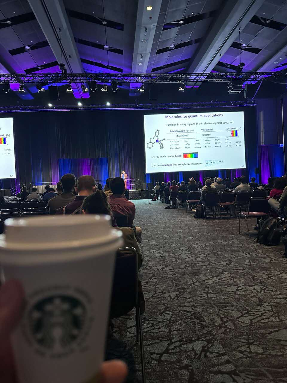 A speaker presenting to a large audience in a conference hall, with a coffee cup in the foreground