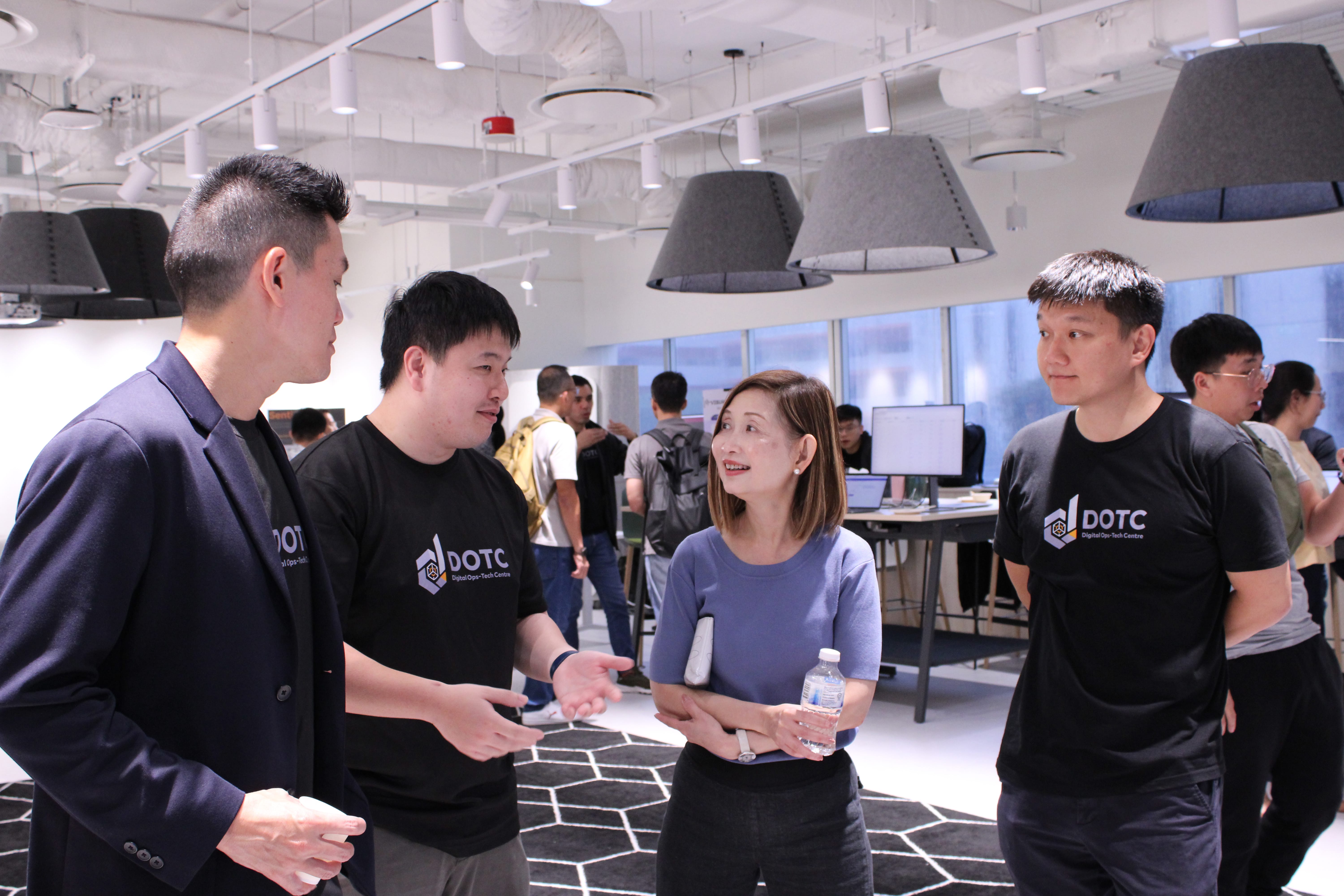 Four people conversing in a modern office, two wearing black DOTC branded t-shirts, with others visible in the background.