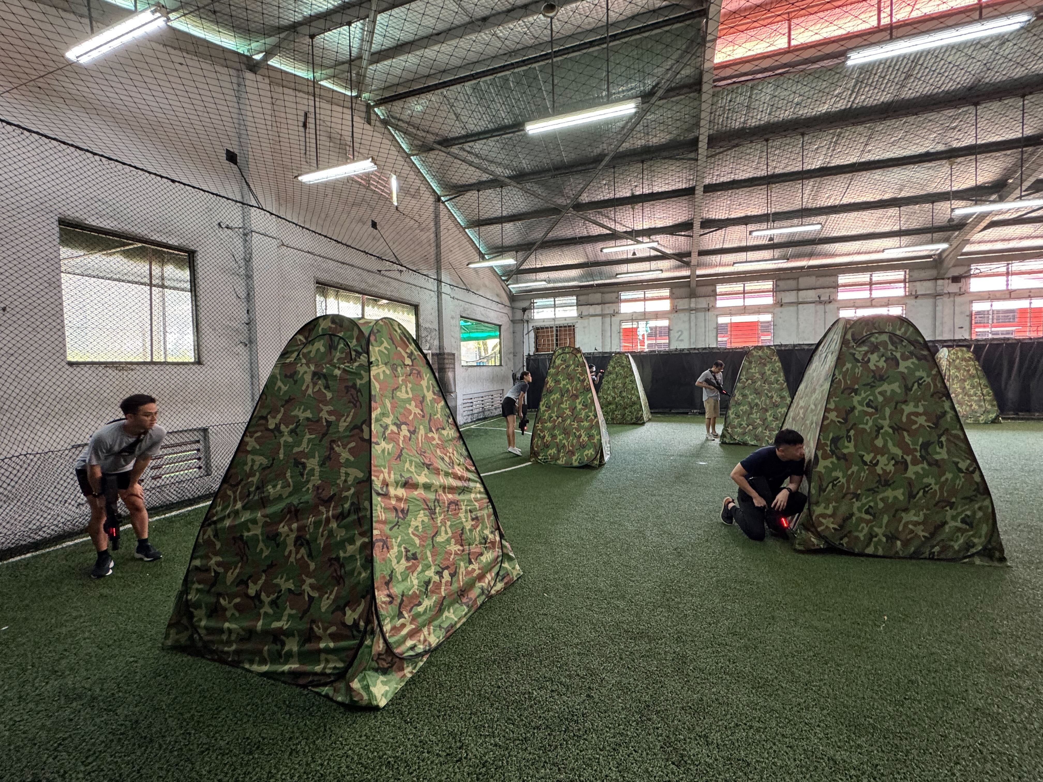 Several camouflage pop-up tents are set up on indoor artificial turf as people crouch and move around them.