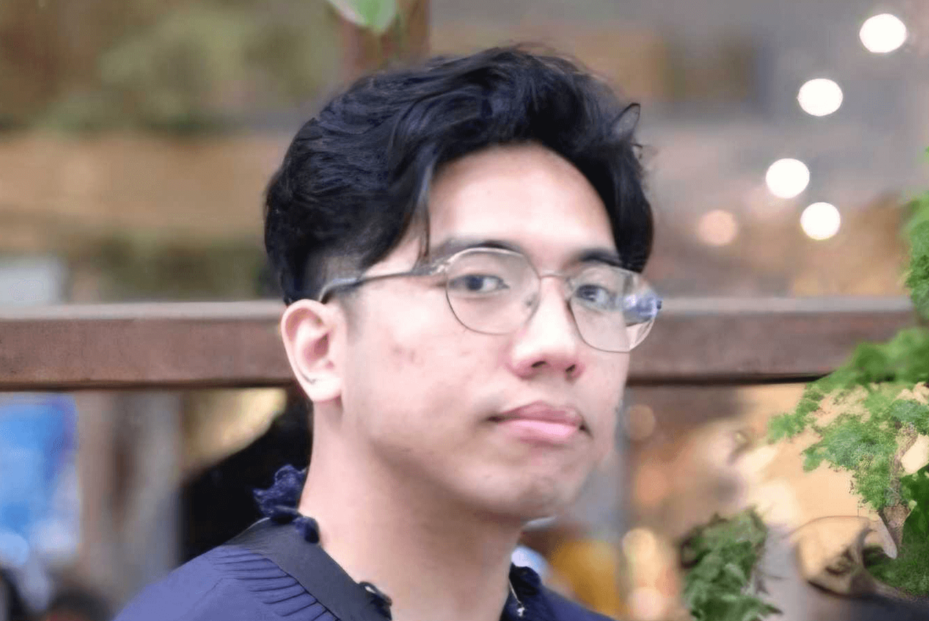 Young man with black wavy hair and clear-framed glasses wearing a navy sweater with a bag strap across his chest.