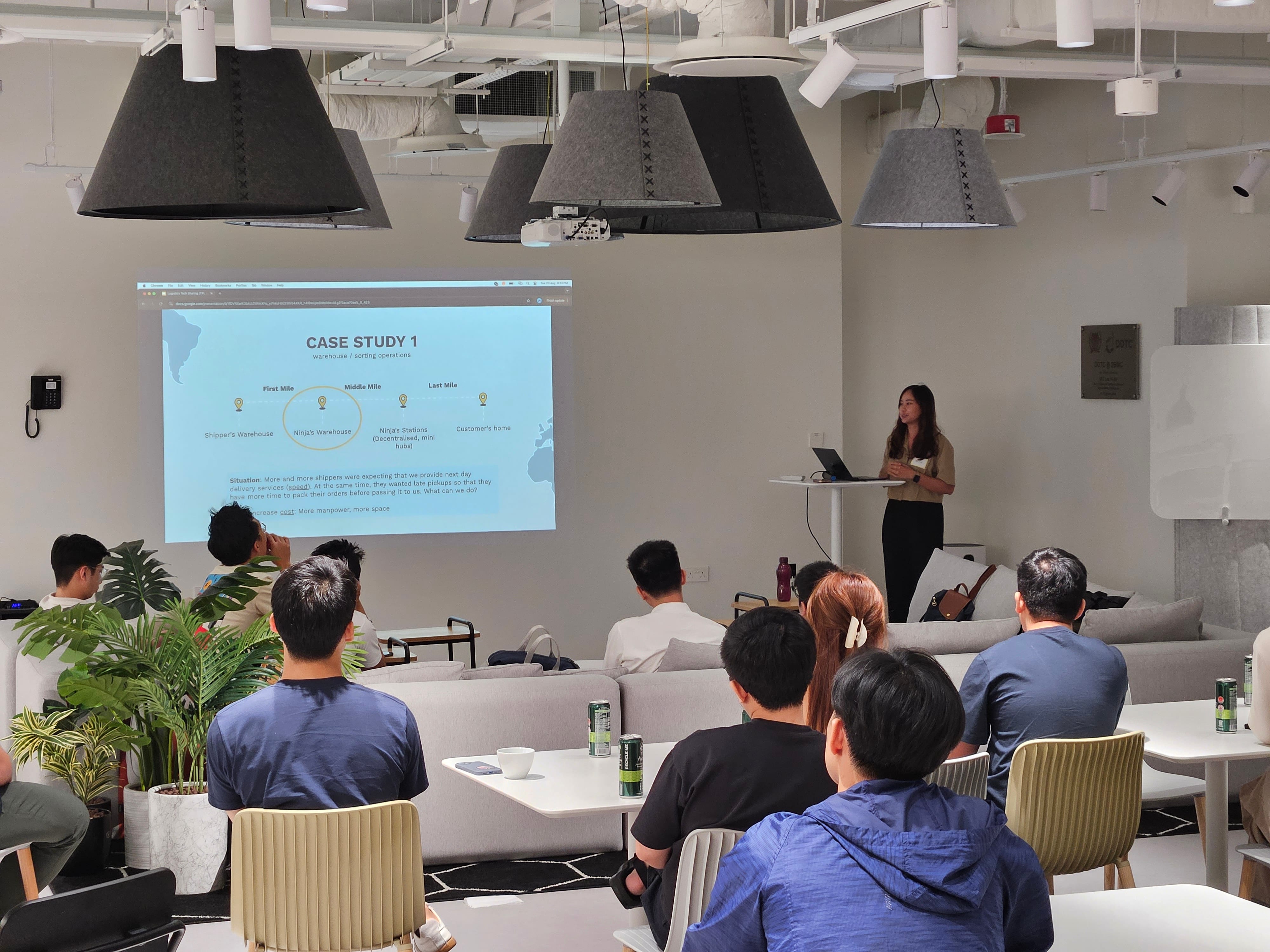 A woman presents a Case Study 1 slide to a seated audience in a modern office lounge with large pendant lamps.